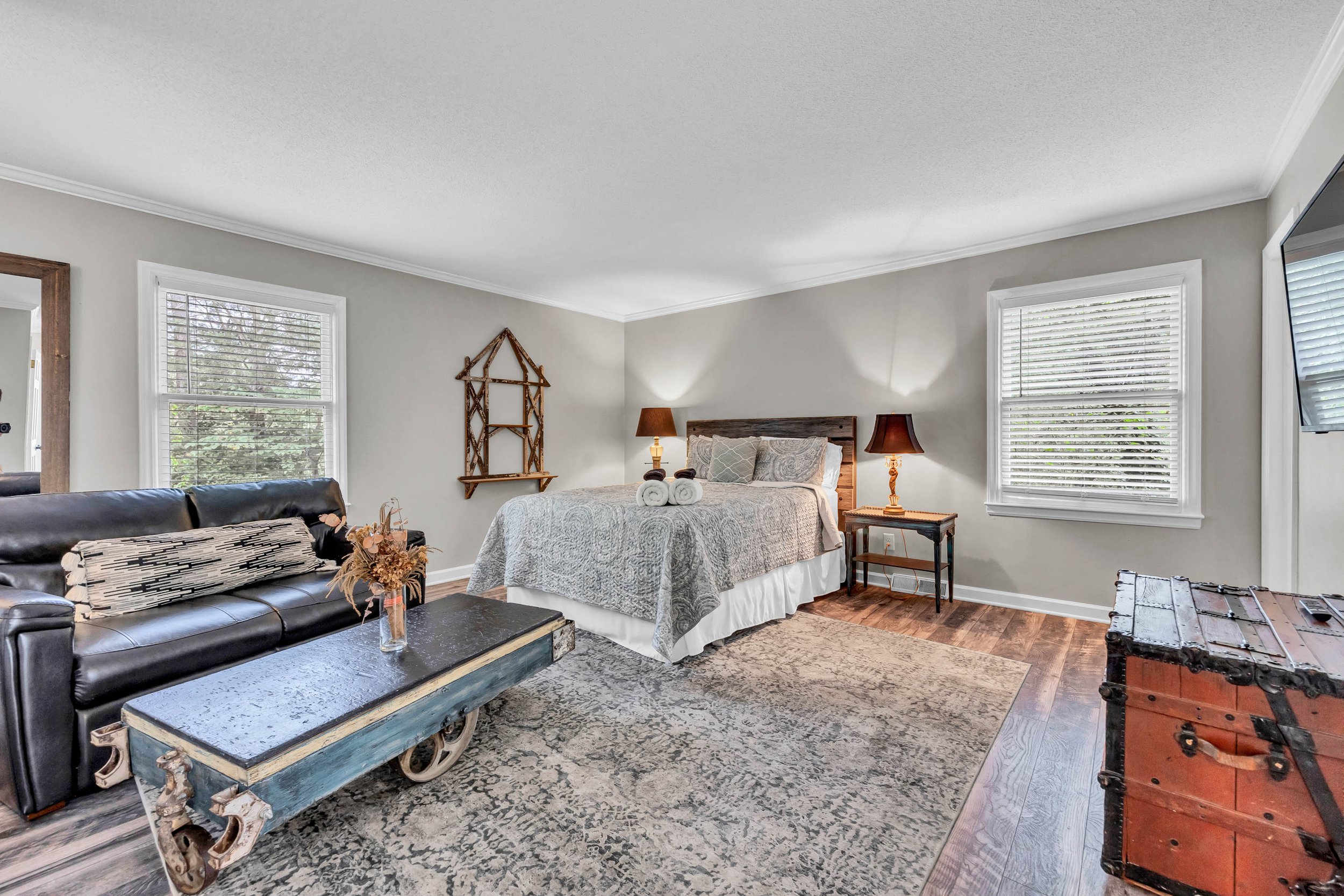 A cozy bedroom with two windows, a bed with a gray patterned quilt, a bedside table with a lamp, a wooden house-shaped wall shelf, a black leather sofa with patterned pillows, a wooden trunk, and a vintage-style coffee table with floral decor."