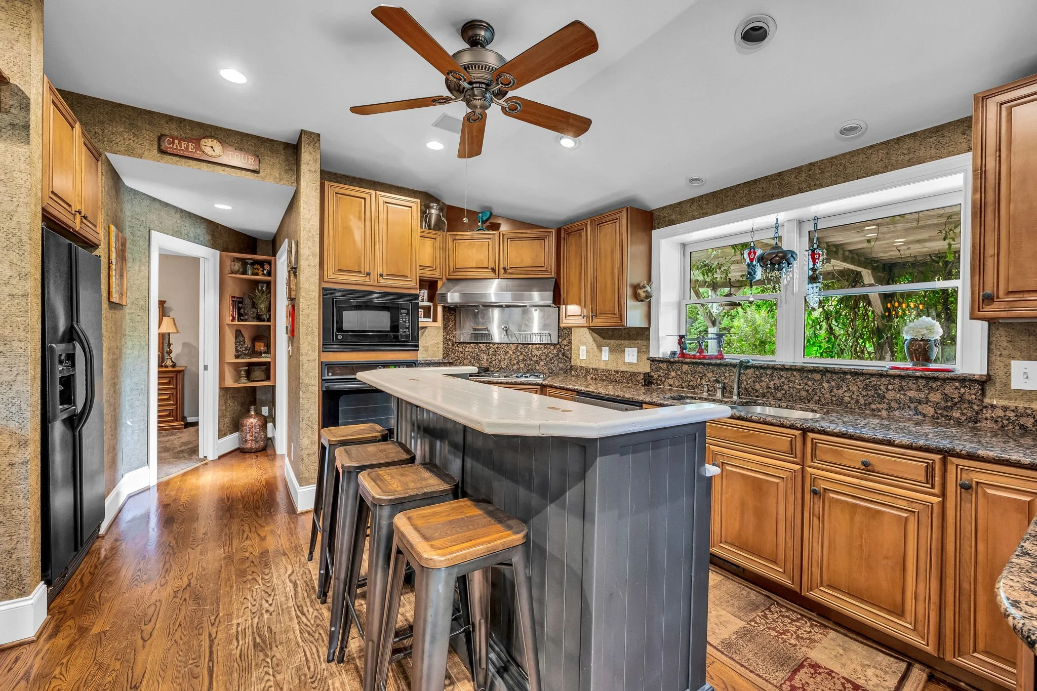 A kitchen with wooden cabinets, a granite countertop, a black refrigerator, a built-in microwave oven, a island with bar stools, a window with hanging decorations, and a ceiling fan.