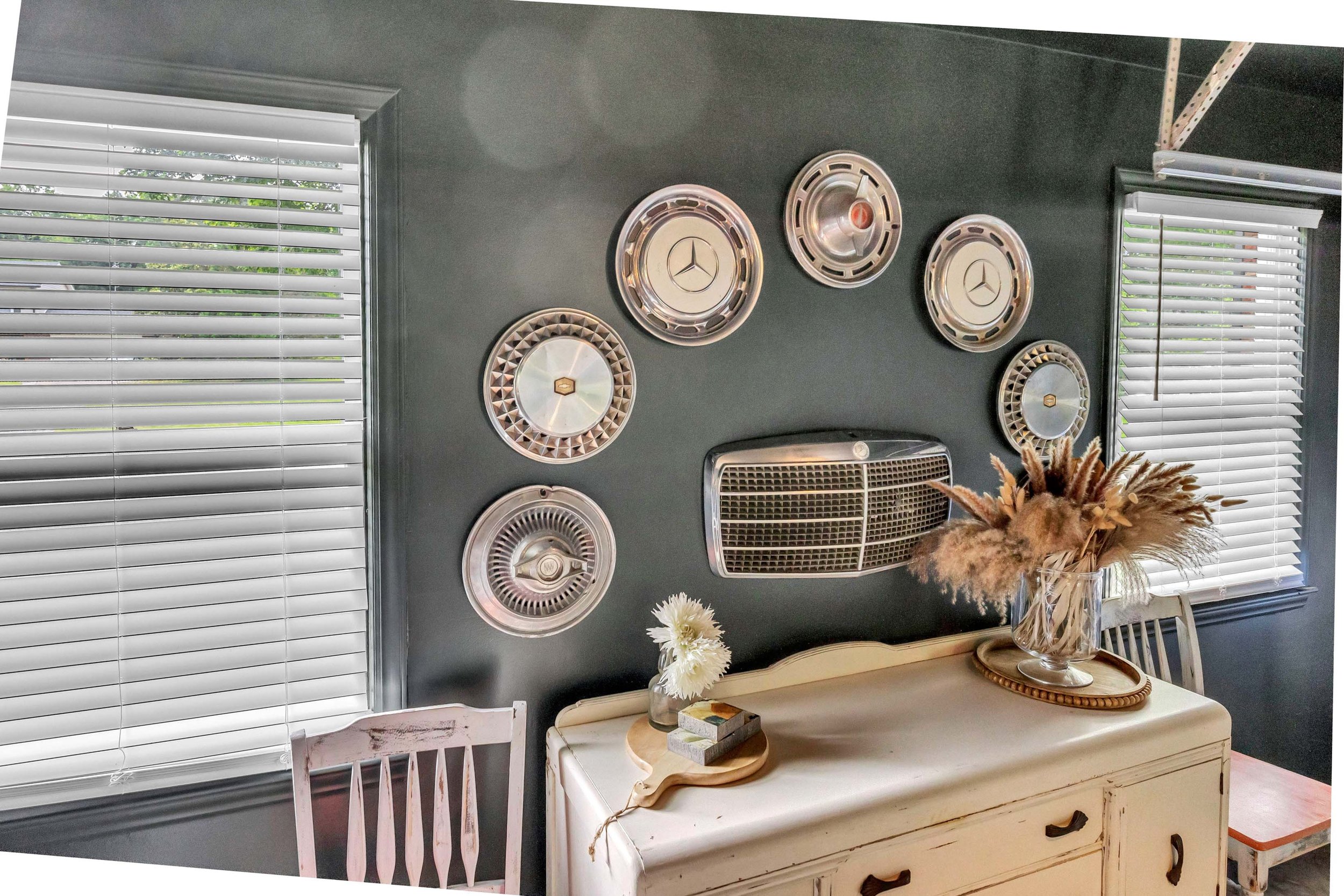A wall decorated with vintage Mercedes-Benz hubcaps, two windows with white blinds, and a table with a vase of dried flowers and a tray with books, in a cozy room with dark walls.