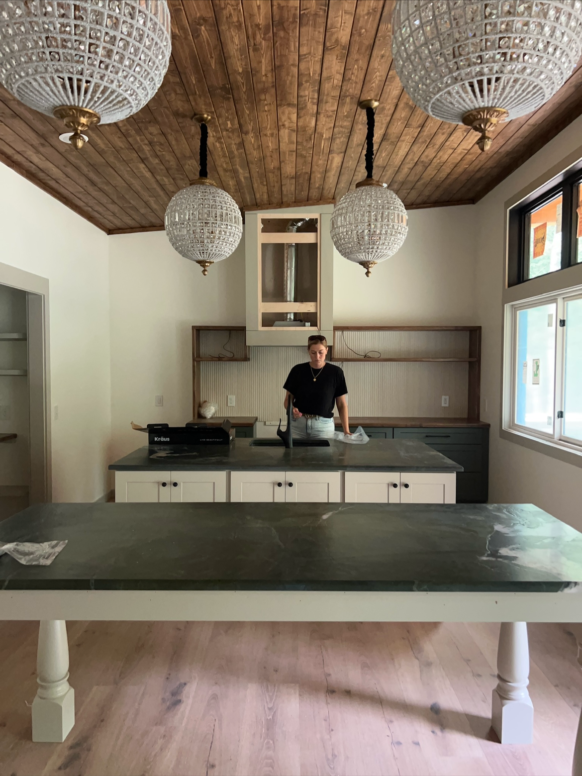 A woman standing in a kitchen with a black shirt and light jeans, surrounded by white cabinets, a wall shelf, a window, and three decorative chandeliers hanging from a wooden ceiling.