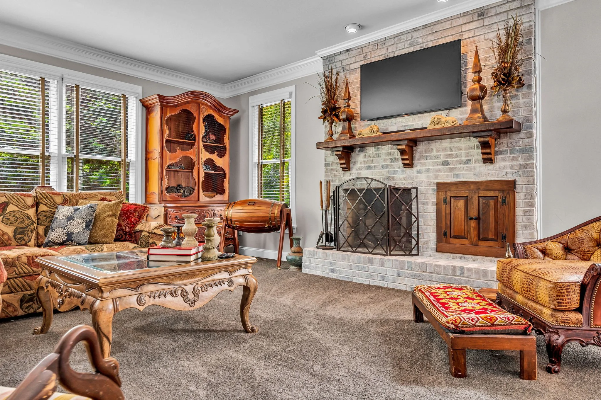 Living room with a brick fireplace, mounted flat-screen TV, wooden mantel, decorative vases, antique-style furniture, patterned sofa with cushions, coffee table, and large windows with blinds.