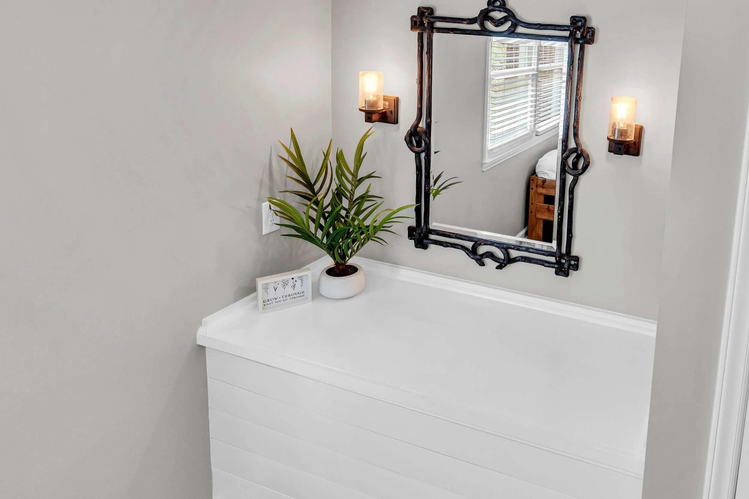 A white dresser with a potted plant, a framed quote, and a decorative mirror with a black, wrought-iron frame. Two wall-mounted light fixtures are on either side of the mirror, and a window with white blinds is reflected in the mirror.