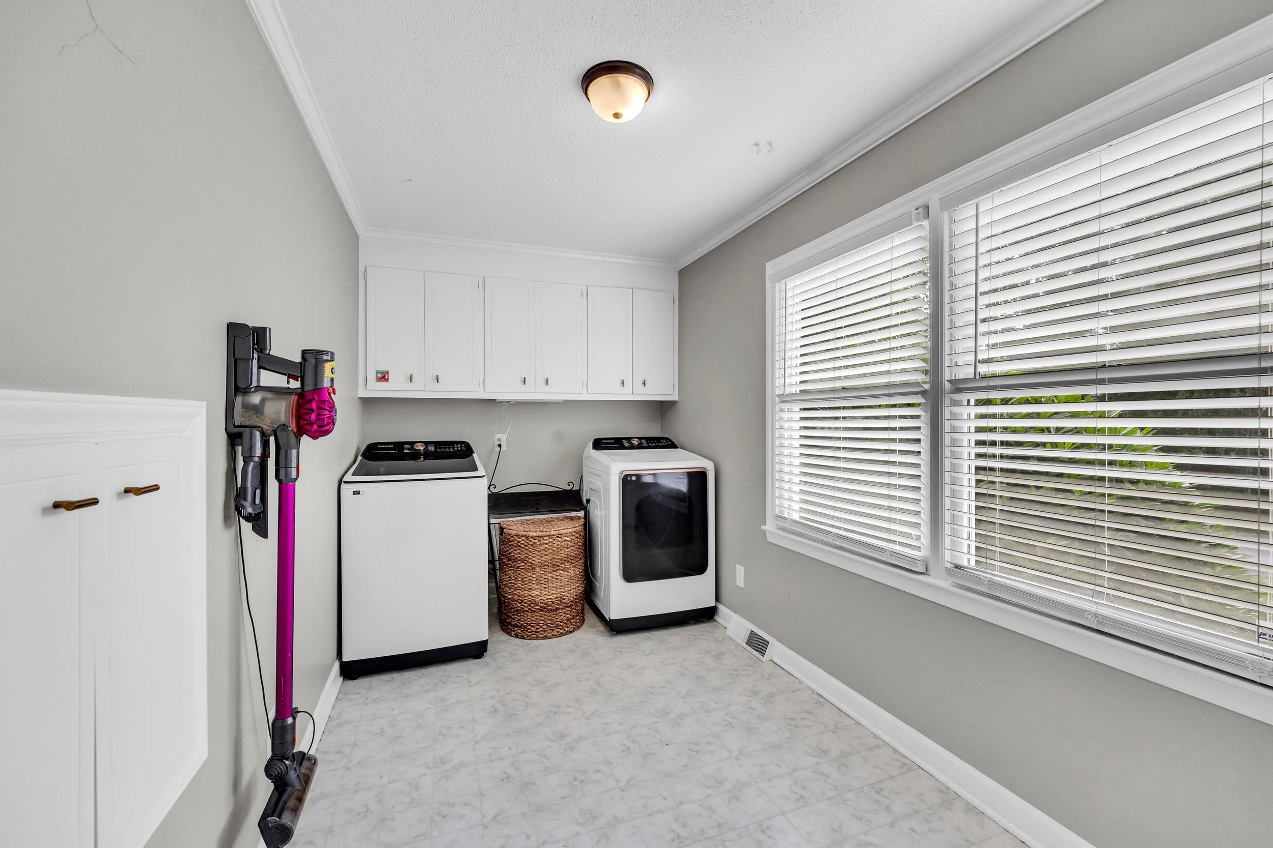 Laundry room with washing machine, dryer, white cabinets, large window with blinds, and a wall-mounted vacuum cleaner.