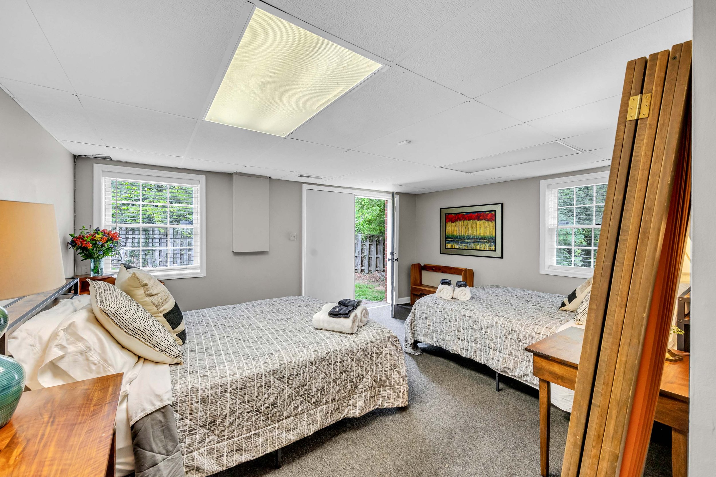A bedroom with two twin beds, each with rolled towels on top, a wooden bench, a framed artwork of flowers on the wall, windows with blinds showing greenery outside, a door leading outside, and a wooden coat rack.