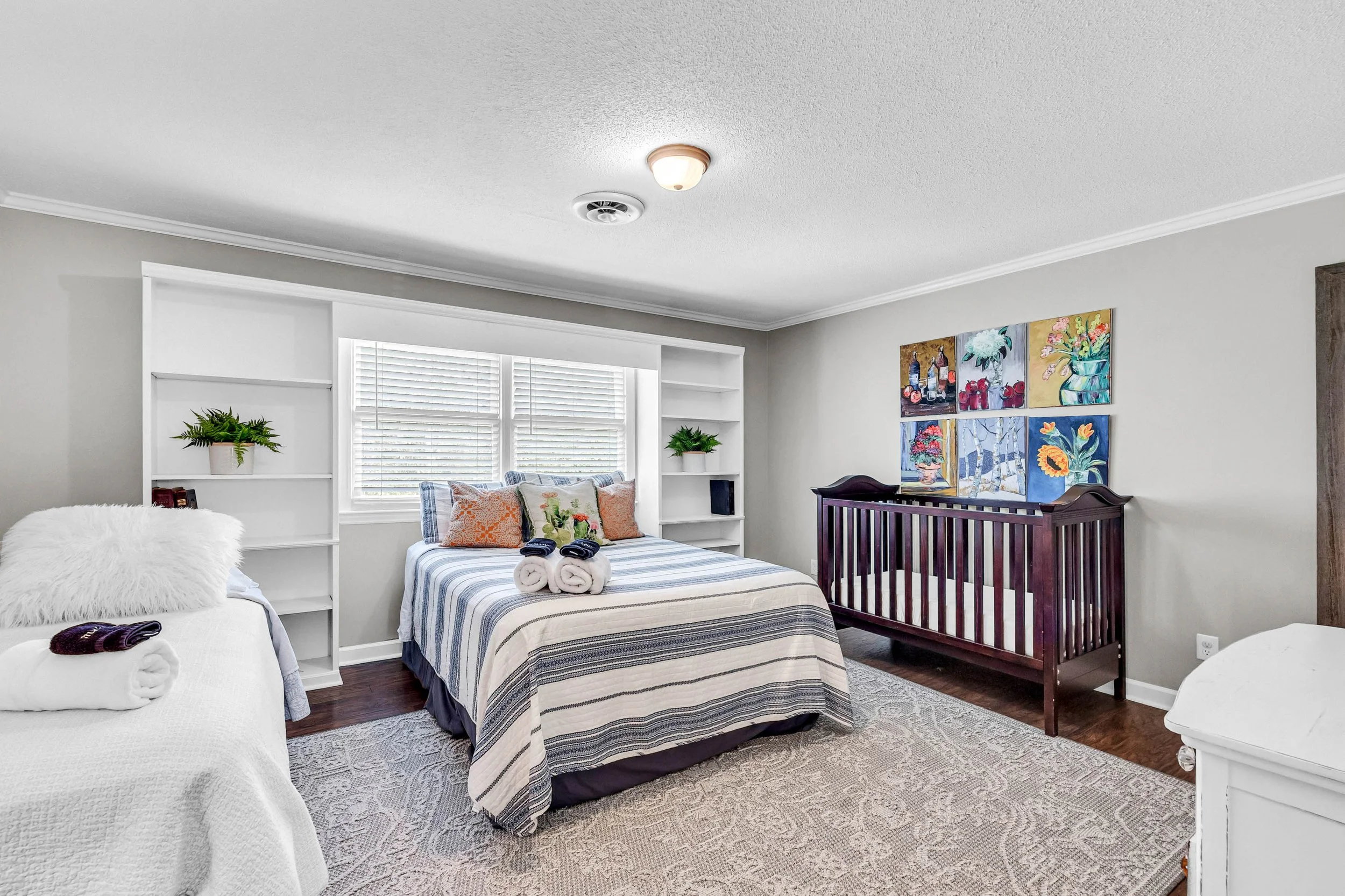 A cozy bedroom with a bed at the center, layered with striped bedding and decorated with various pillows. There's a window with white blinds behind the bed, allowing natural light in. The walls are painted light gray, with colorful paintings hanging on one wall. To the right, a dark wooden baby crib is visible. On the left, a white dresser and shelves with green plants and books are present. The ceiling has a light fixture and a smoke detector.
