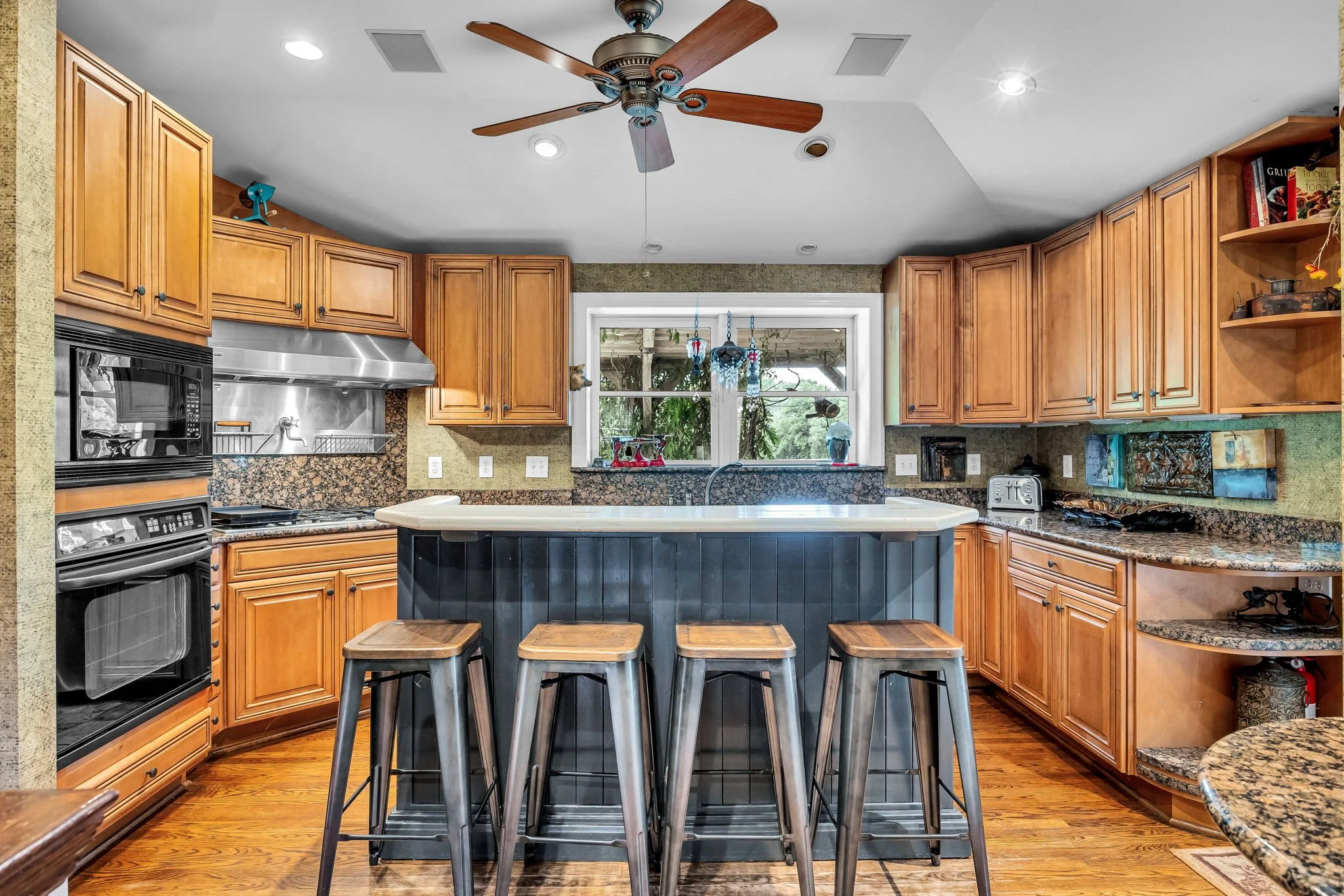 A kitchen with wooden cabinets, granite countertops, stainless steel appliances, and a window above the sink. There is a kitchen island with four stools, ceiling fan, and various decorative items and kitchen accessories.