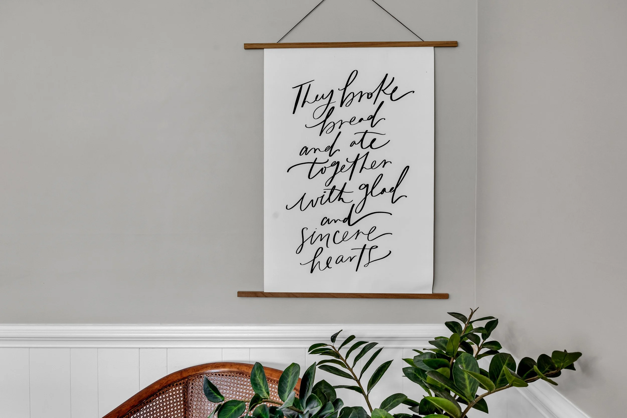 A decorative wall hanging with a written message hangs above a white wainscoting wall. The message reads: "They broke bread and ate together with glad and sincere hearts." The wall hanging is supported by wooden rods at the top and bottom, and is suspended by a thin string. In the lower part of the image, a green leafy plant and a wooden chair back are visible.