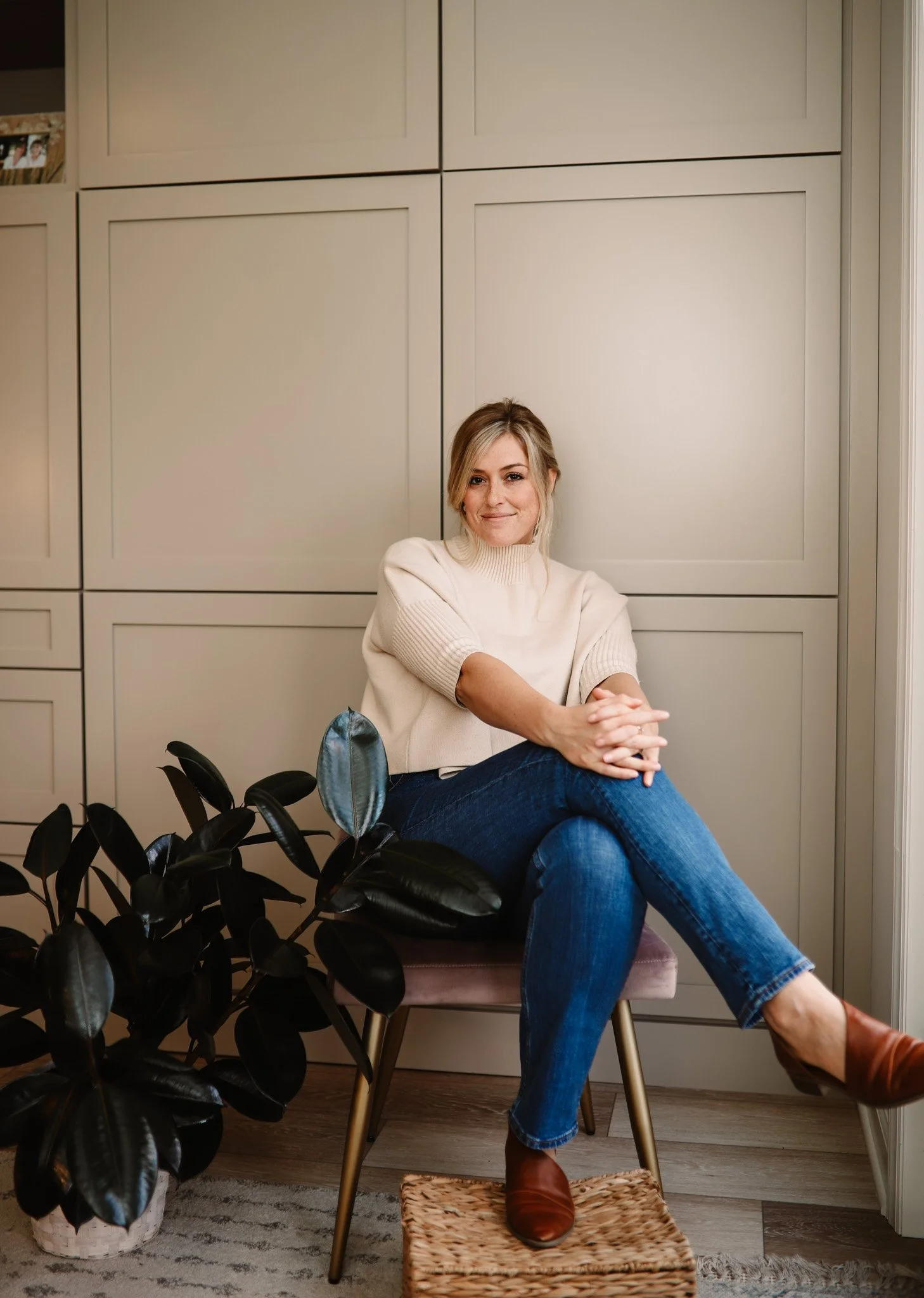 A woman with blonde hair in a beige sweater and blue jeans sitting on a chair in front of a beige cabinet, with one foot on a wicker footstool and the other resting on the floor. She is smiling and looking at the camera.