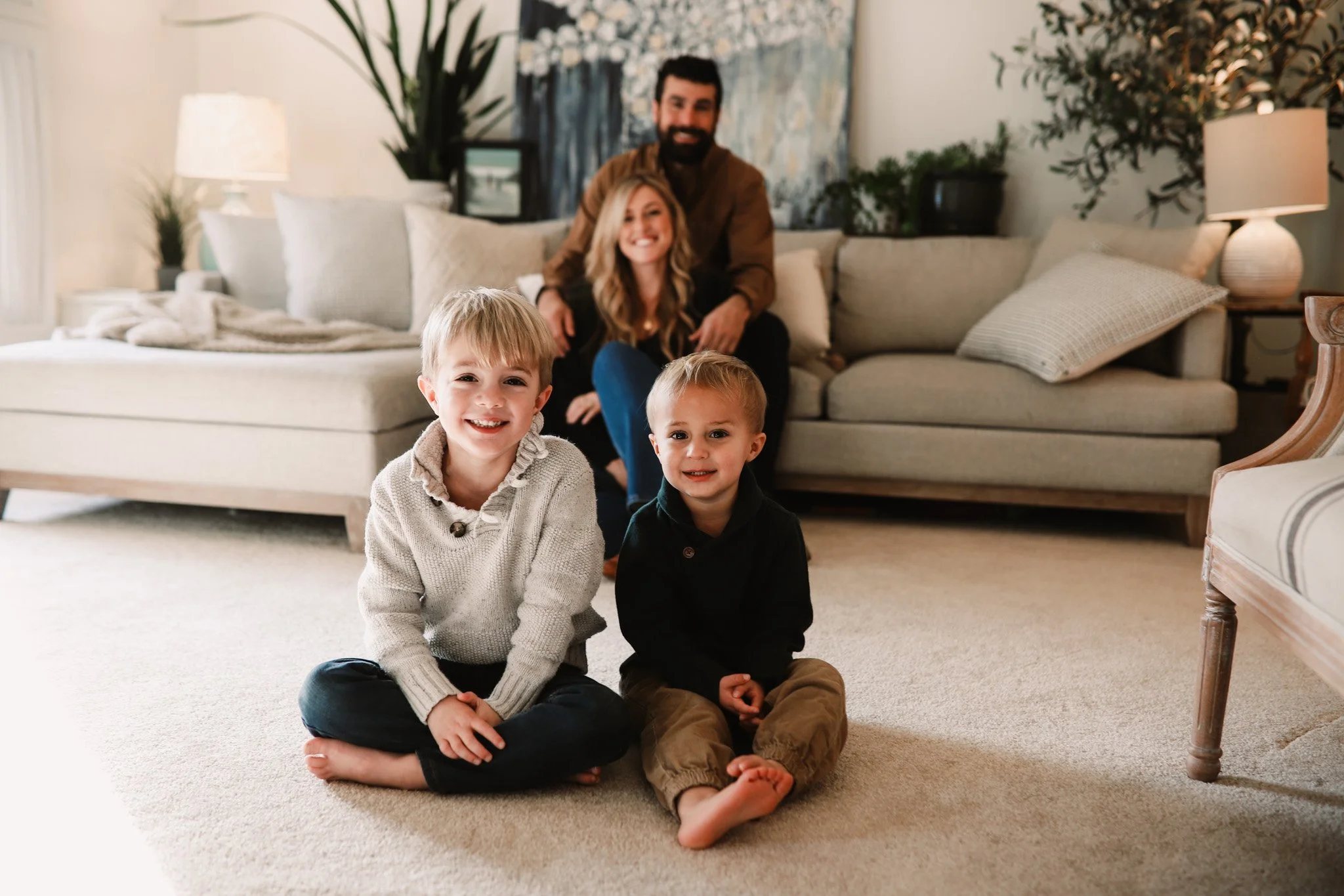 Two young boys sitting on the floor with a happy family of four in the blurred background inside a living room with a beige couch, pillows, and plants.