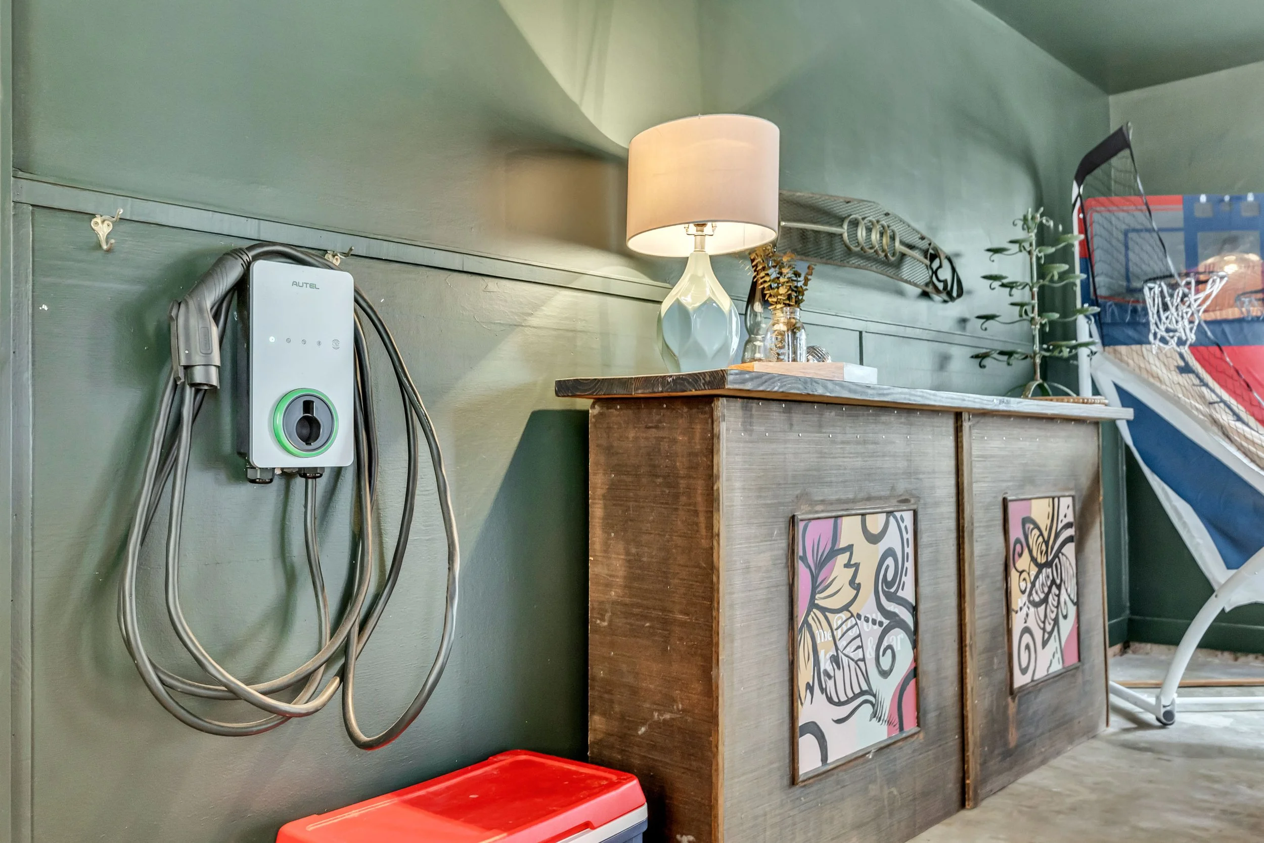 The image shows a green wall with various decorative items and furniture. There is a white lamp with a beige shade, a glass jar with a plant, a wooden cabinet with artwork, and an art piece, as well as an electrical device with cords plugged into it on the left side.