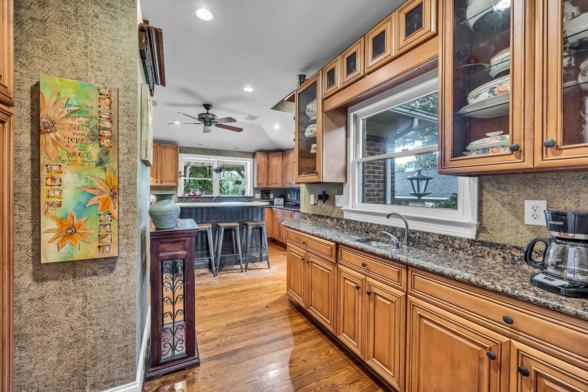 View of a kitchen with wooden cabinets, granite countertops, a window above the sink, and decorative plates in glass cabinets. There is a colorful wall art with a motivational quote on the left side and a ceiling fan in the background.