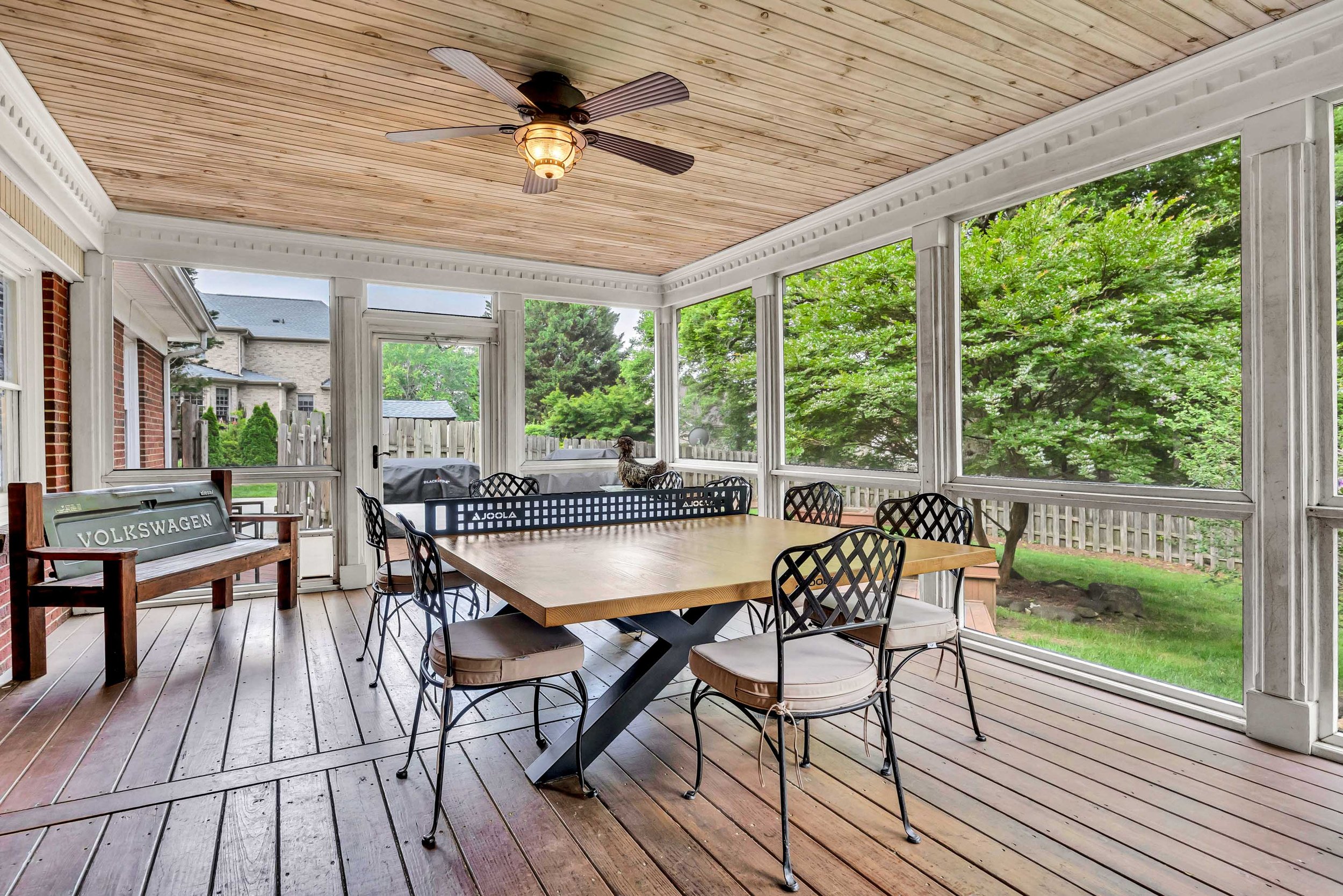 Enclosed porch with wooden ceiling, ceiling fan, large wooden table, black metal chairs, wooden bench with Volkswagen logo, and view of green trees and neighboring houses outside.