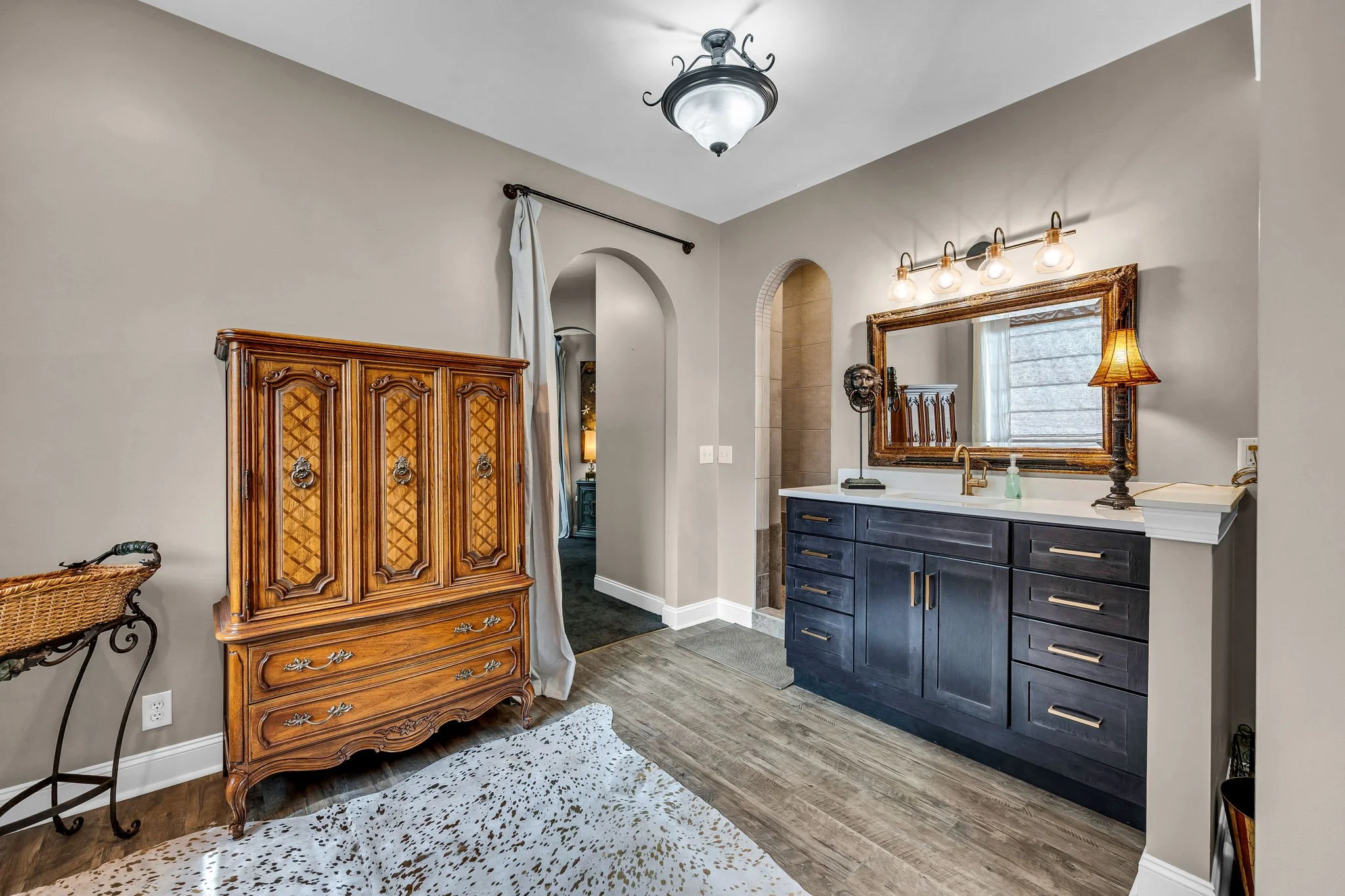 A bathroom with a navy blue vanity, large mirror, light fixture with four bulbs, decorative lamp, and an antique wooden cabinet.