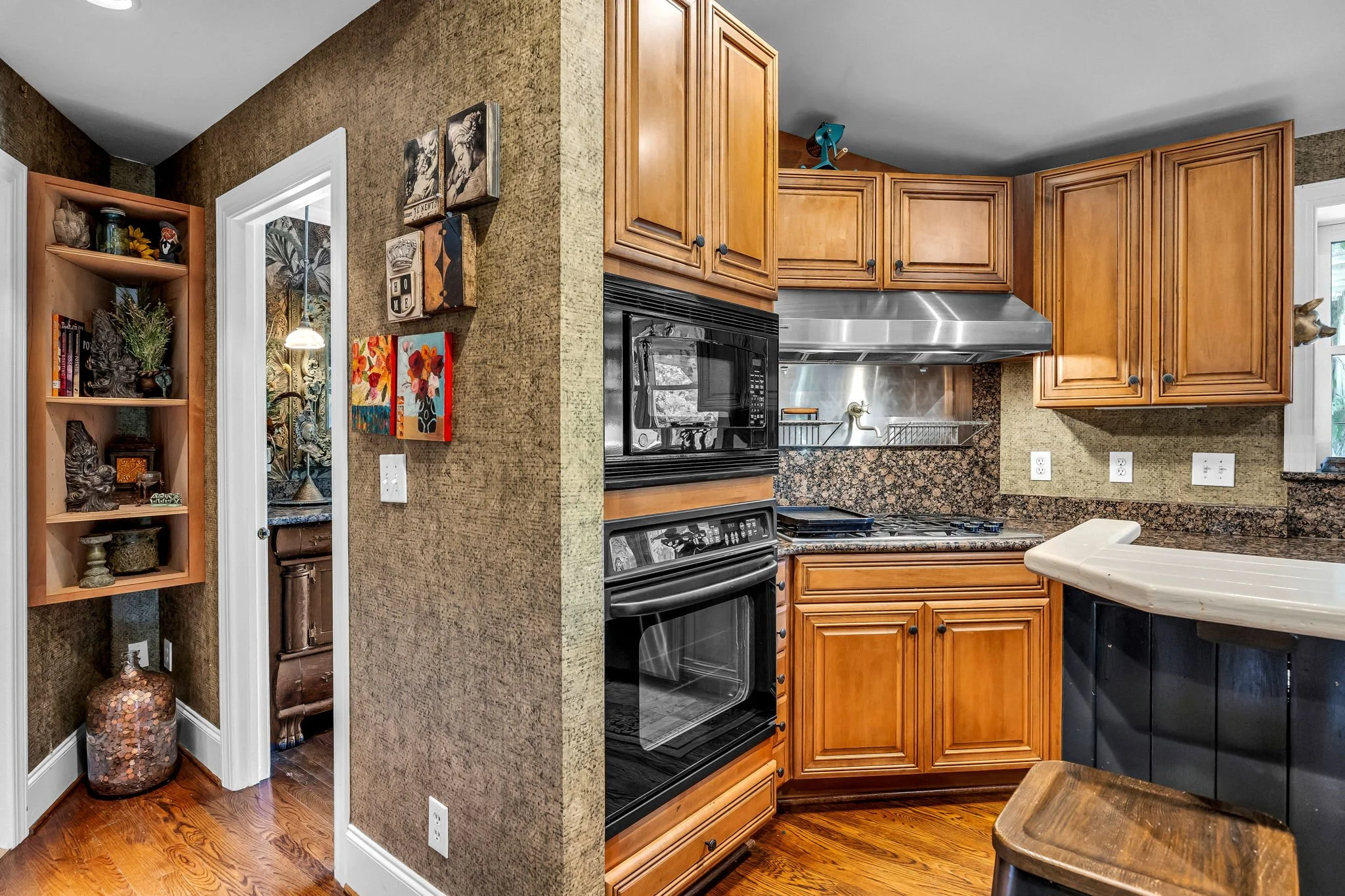 Kitchen with wooden cabinets, black oven, stainless steel microwave, granite countertops, and a small table with a wooden stool. Decorative items on a shelf and artwork on the wall are also visible.