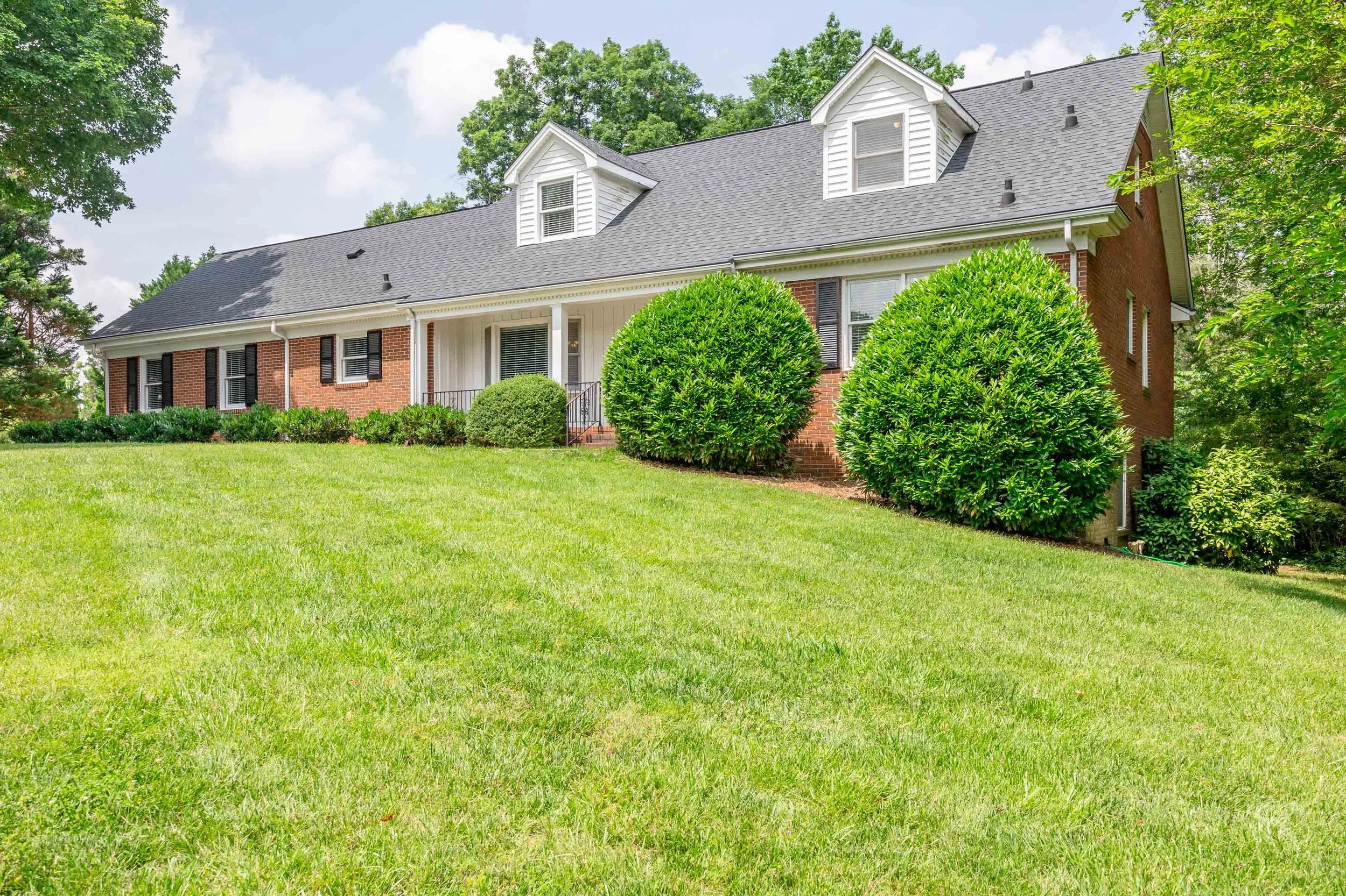 Front view of a large house with red brick walls, white siding, a dark gray shingle roof, and two dormer windows. The house is surrounded by well-maintained green bushes and trees, with a lush green lawn in the foreground and blue sky with some clouds above.