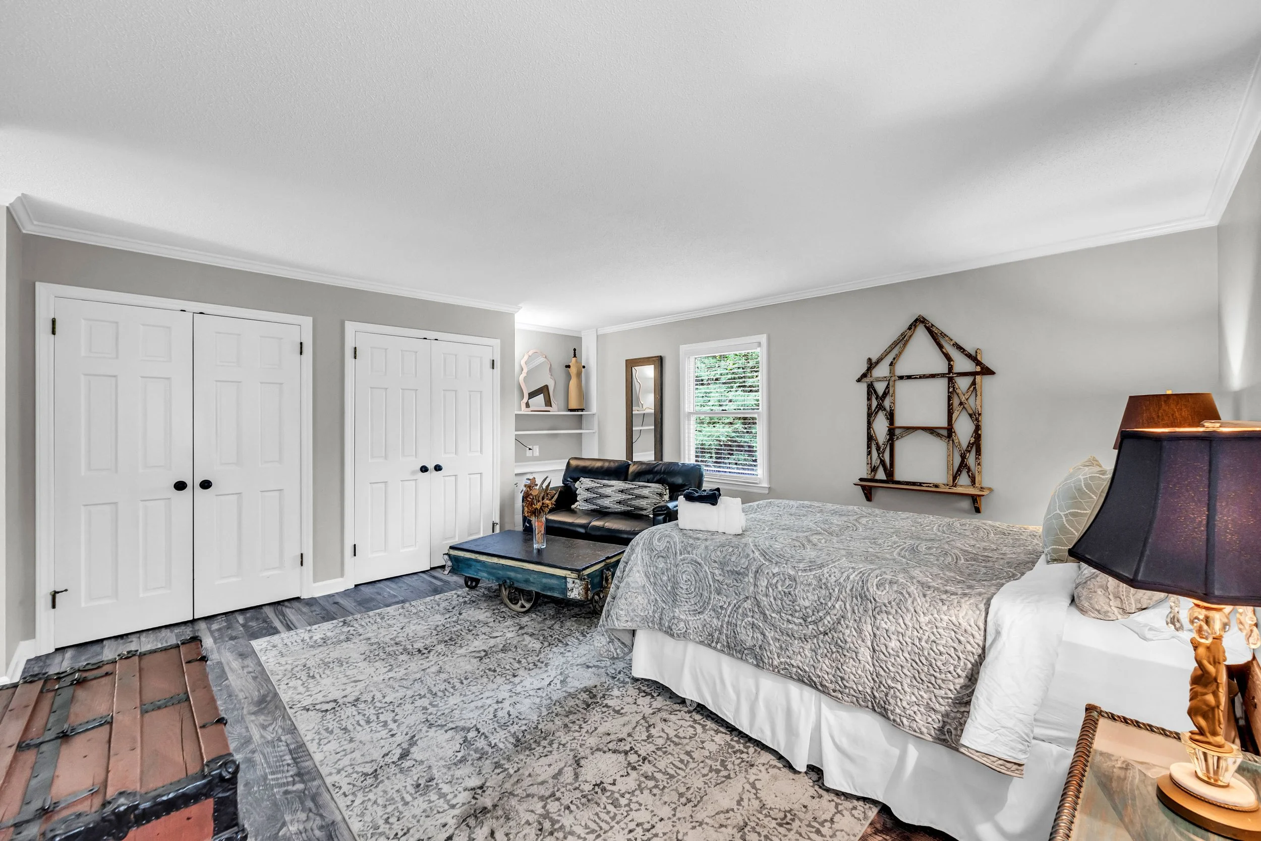 A bedroom with a gray and white color scheme, including a bed with a patterned bedspread, a black leather chair, possibly a coffee table, and decorative items on the walls. There are white double closet doors and a window with blinds, with a mix of modern and rustic decor elements.