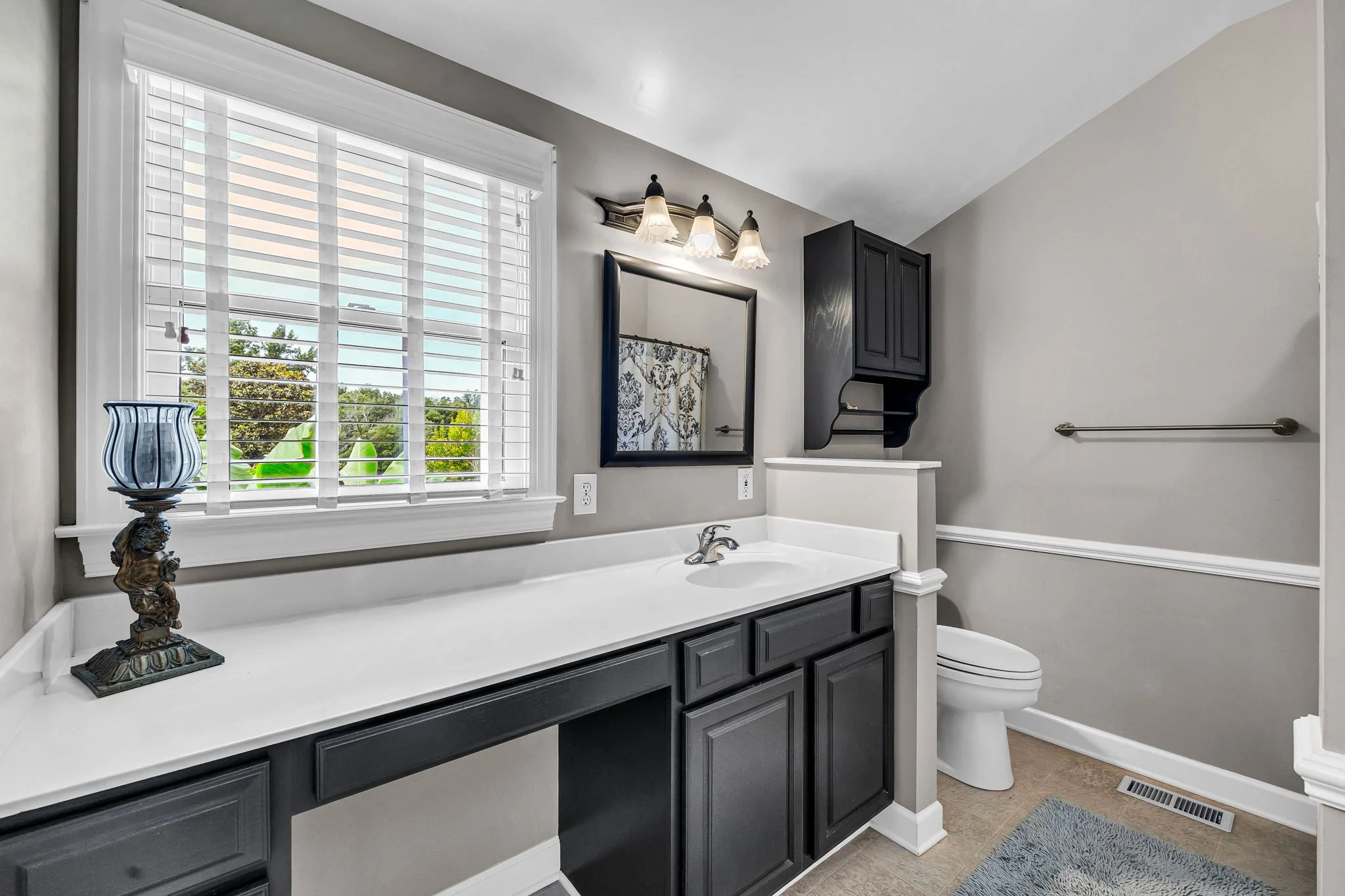 Bathroom with a large window with white blinds, a black vanity with multiple drawers, a mirror, a black wall cabinet, a decorative lamp on a stand, and a toilet. The walls are painted light gray, and there is a blue rug on the beige tiled floor.