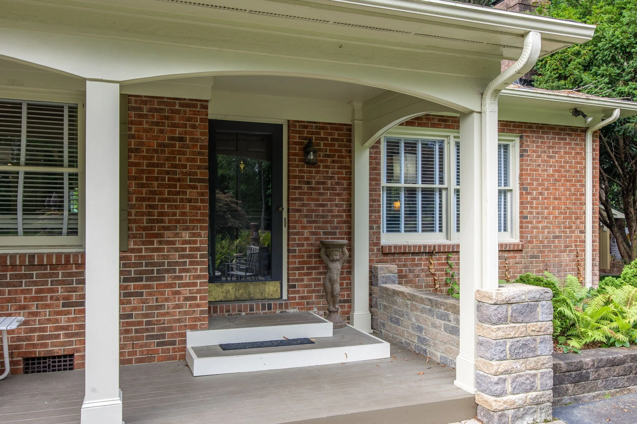 The front porch of a brick house with white trim, a black front door, and a small set of steps leading up to it. There is a decorative stone urn beside the door and a small overhanging roof with a light fixture. The house features large windows with white shutters and is surrounded by greenery.