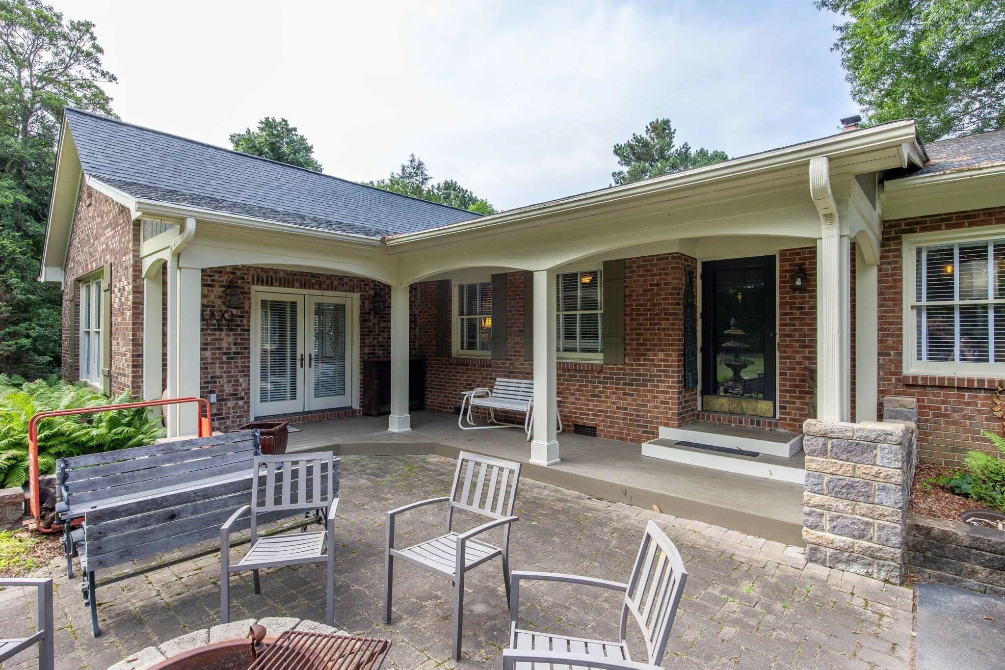 A backyard patio with multiple chairs, a brick house with a covered porch, and a wooded area in the background.