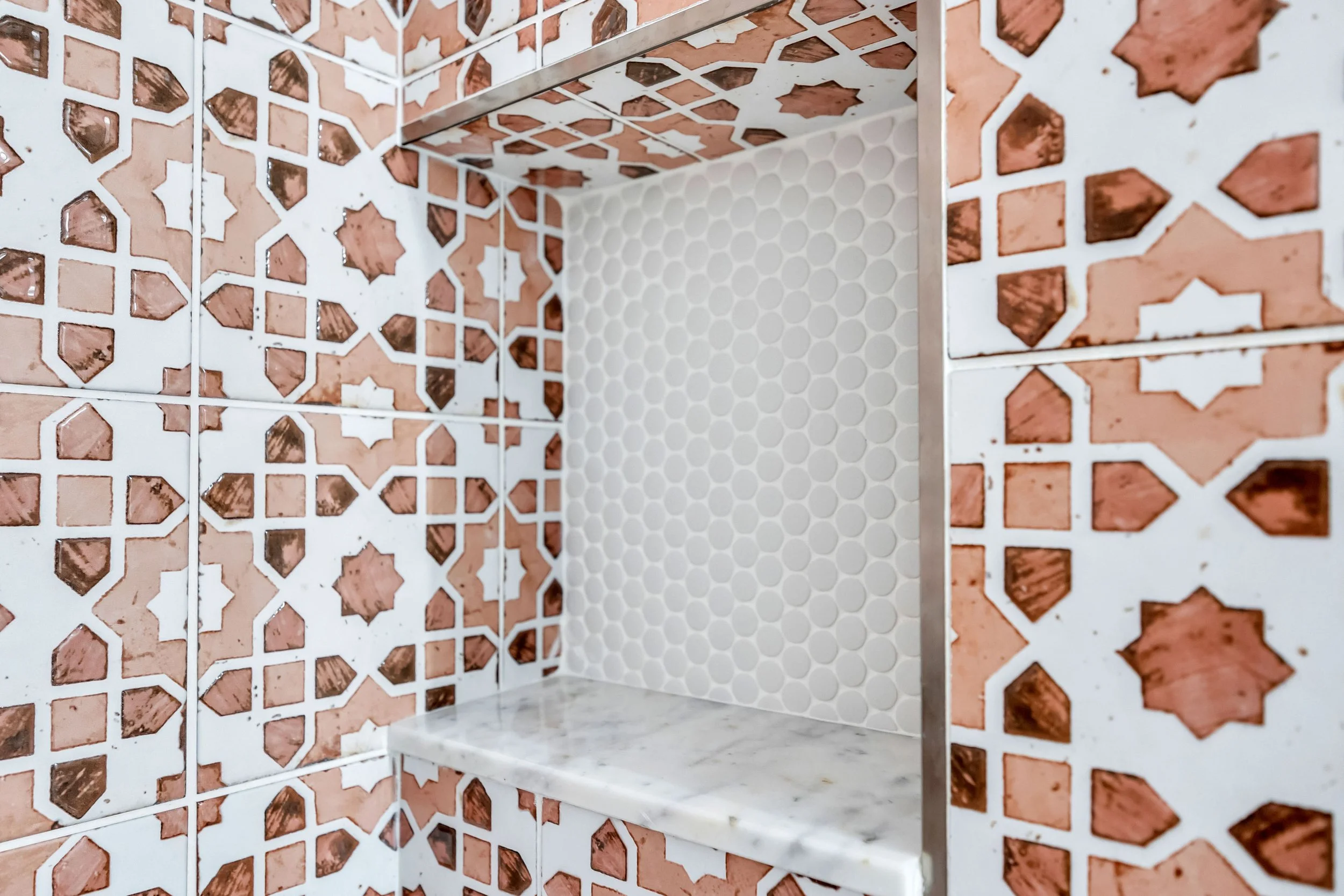 Close-up of a corner of a wall with ornate patterned tiles featuring pinkish-brown star and geometric shapes, and a section of white honeycomb tiles on the wall with a marble ledge at the bottom.