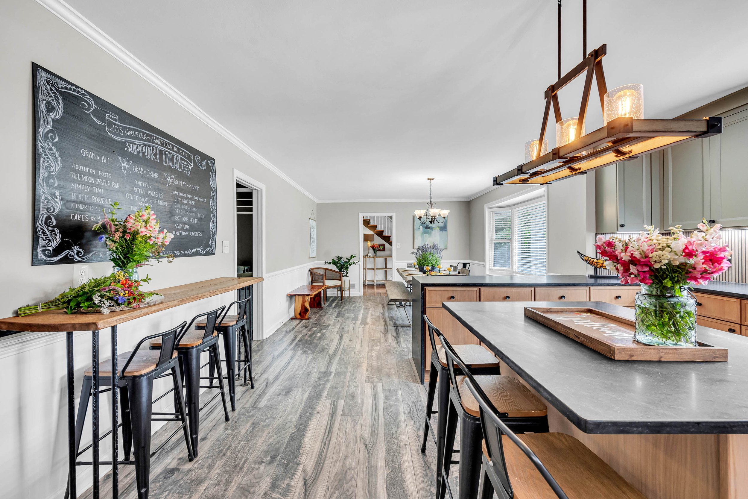 A modern, bright kitchen and dining area with large windows, floating black bar stools at a kitchen island, a chalkboard wall with handwritten menu, floral arrangements, and a rustic chandelier.