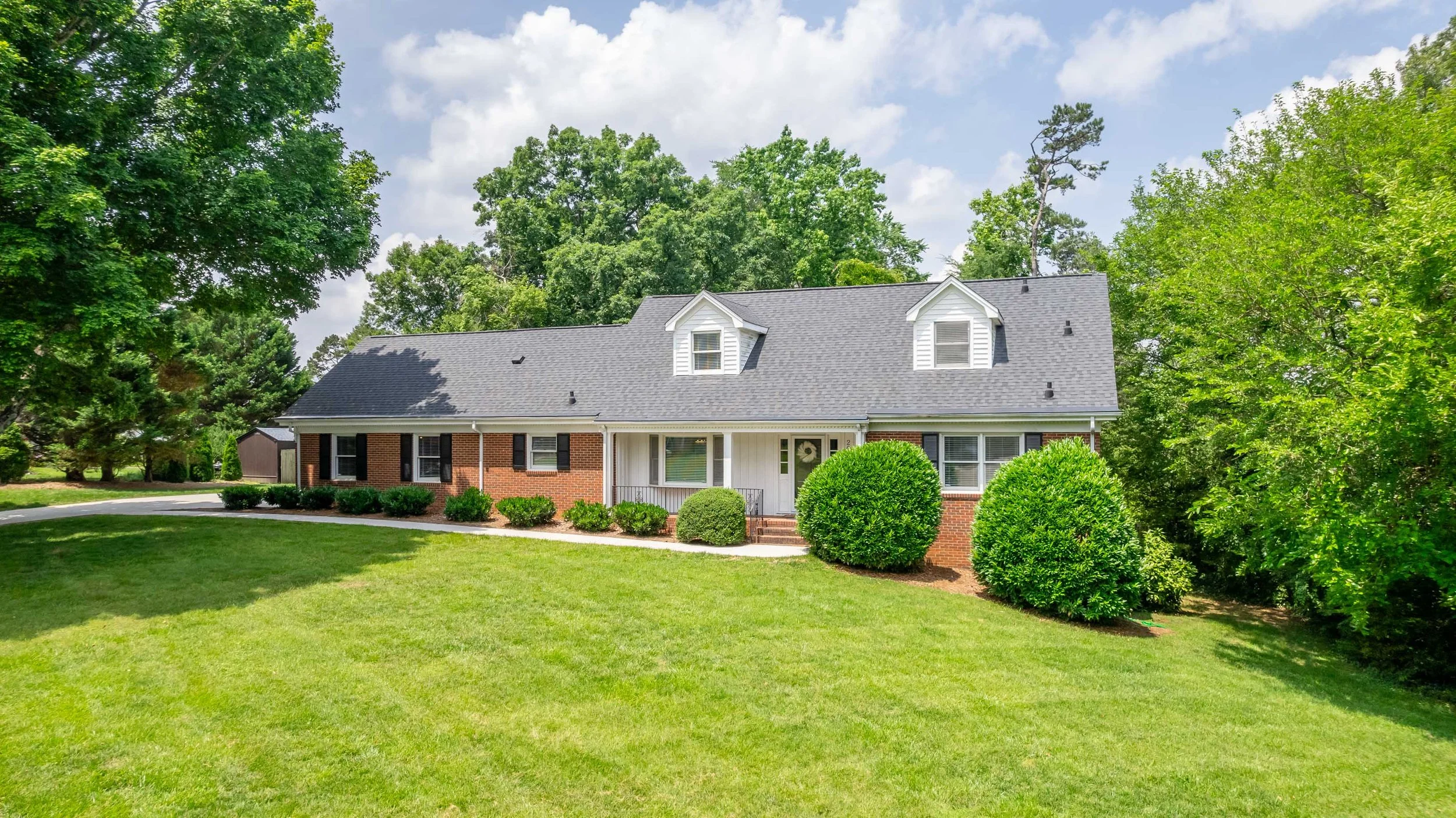 A two-story house with a gray roof, brick and white siding exterior, and black shutters, surrounded by lush green trees and shrubs, with a well-maintained lawn.