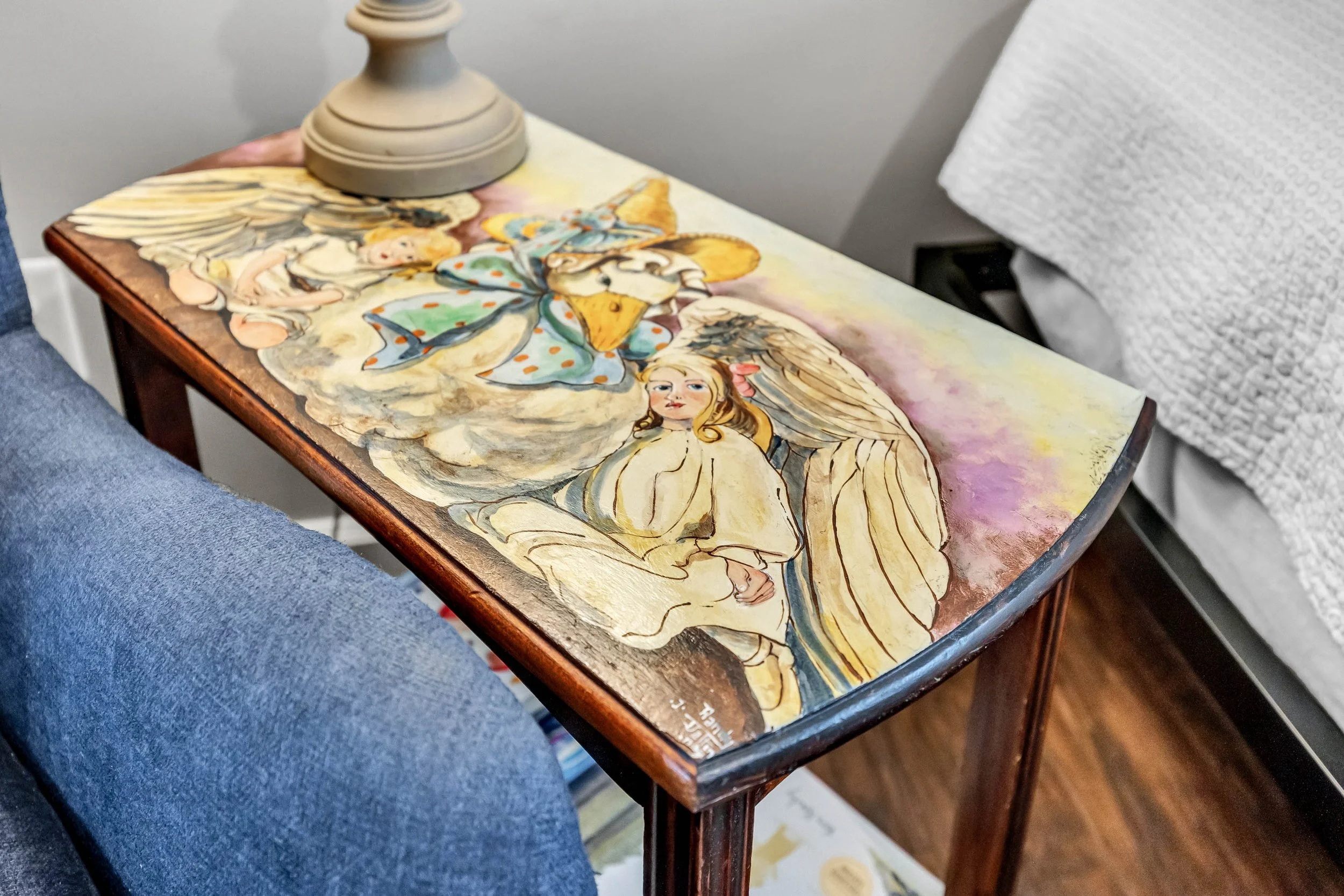 Close-up of a wooden table with a colorful hand-painted illustration of fairies, butterflies, and flowers, topped with a beige lamp base, with a person's jeans-clad knee visible on the left and a white textured cushion in the background.