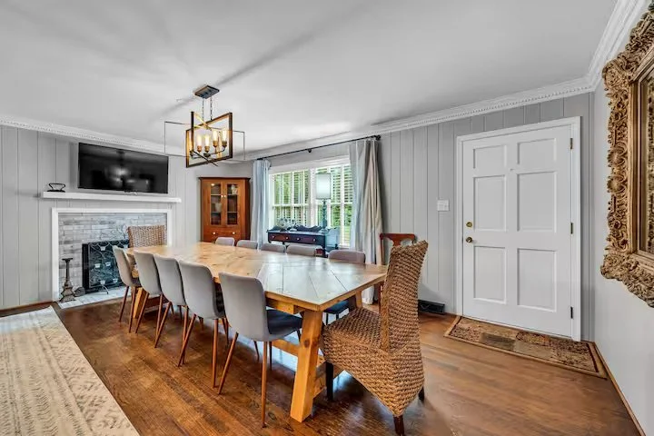 Dining room with a large wooden table, eight chairs, a chandelier, a corner cabinet, a fireplace with a TV above it, and a window with curtains showing outside greenery.