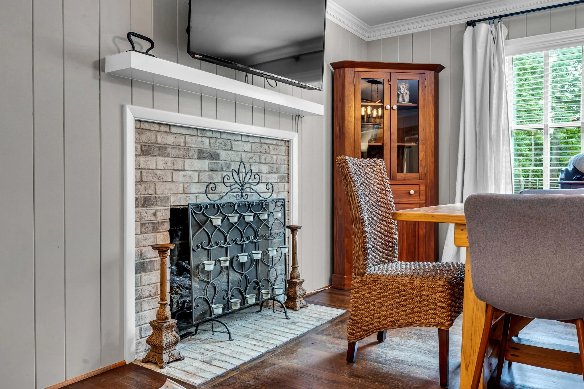 Living room with a brick fireplace with a black metal and white candleholder, a wooden cabinet with glass doors, a woven chair, a wooden dining table, and a window with white curtains showing green trees outside.