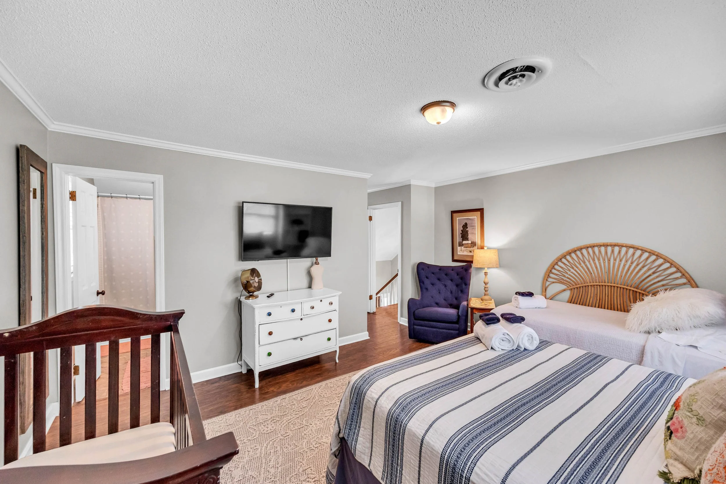 A bedroom with a bed, purple armchair, dresser, wall-mounted TV, framed artwork, and a nightstand with a lamp. Towels are on the bed, and the room has hardwood floors and light-colored walls.