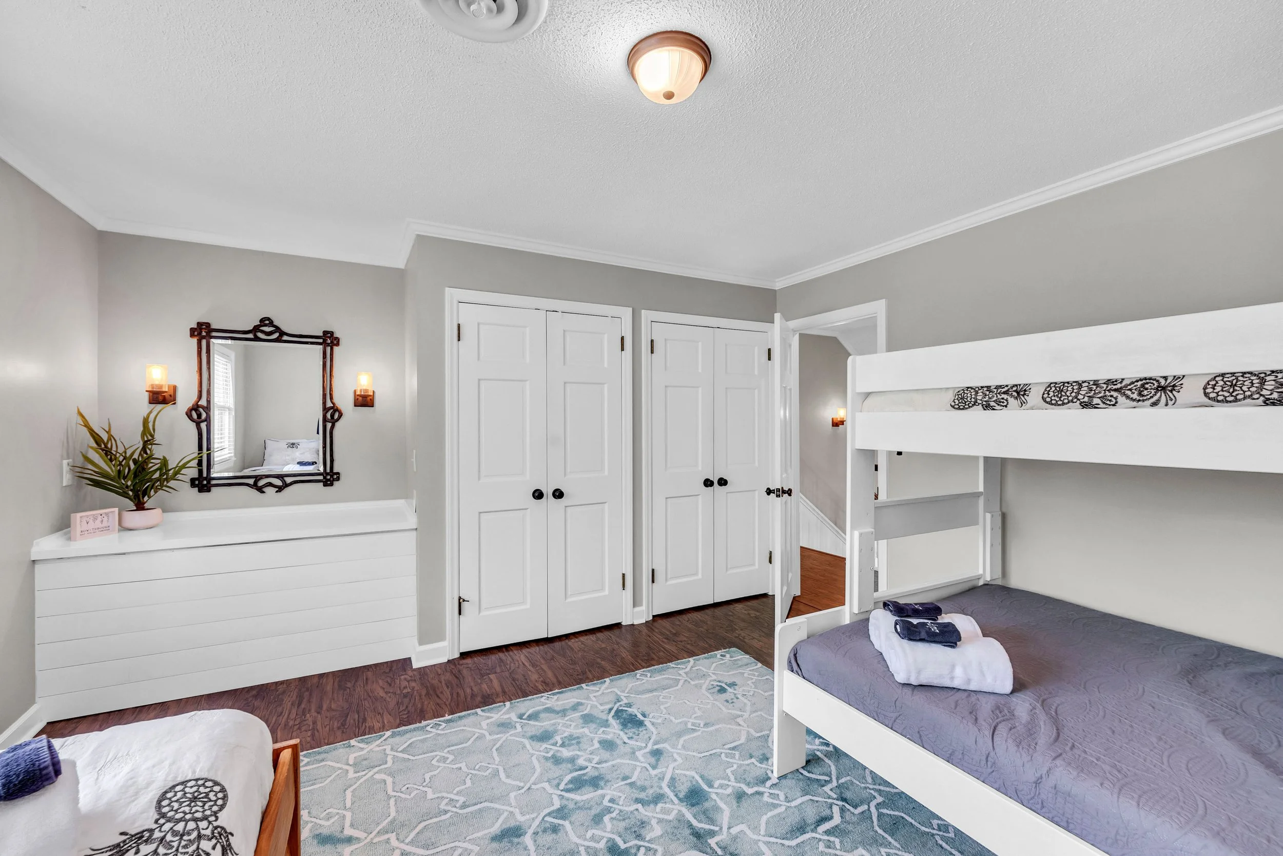 A bedroom with white walls and closet doors, a bunk bed with a purple mattress, a blue and white patterned rug, a mirror with a decorative wooden frame, and a small potted plant. There are two wall-mounted lamps and a ceiling light fixture.