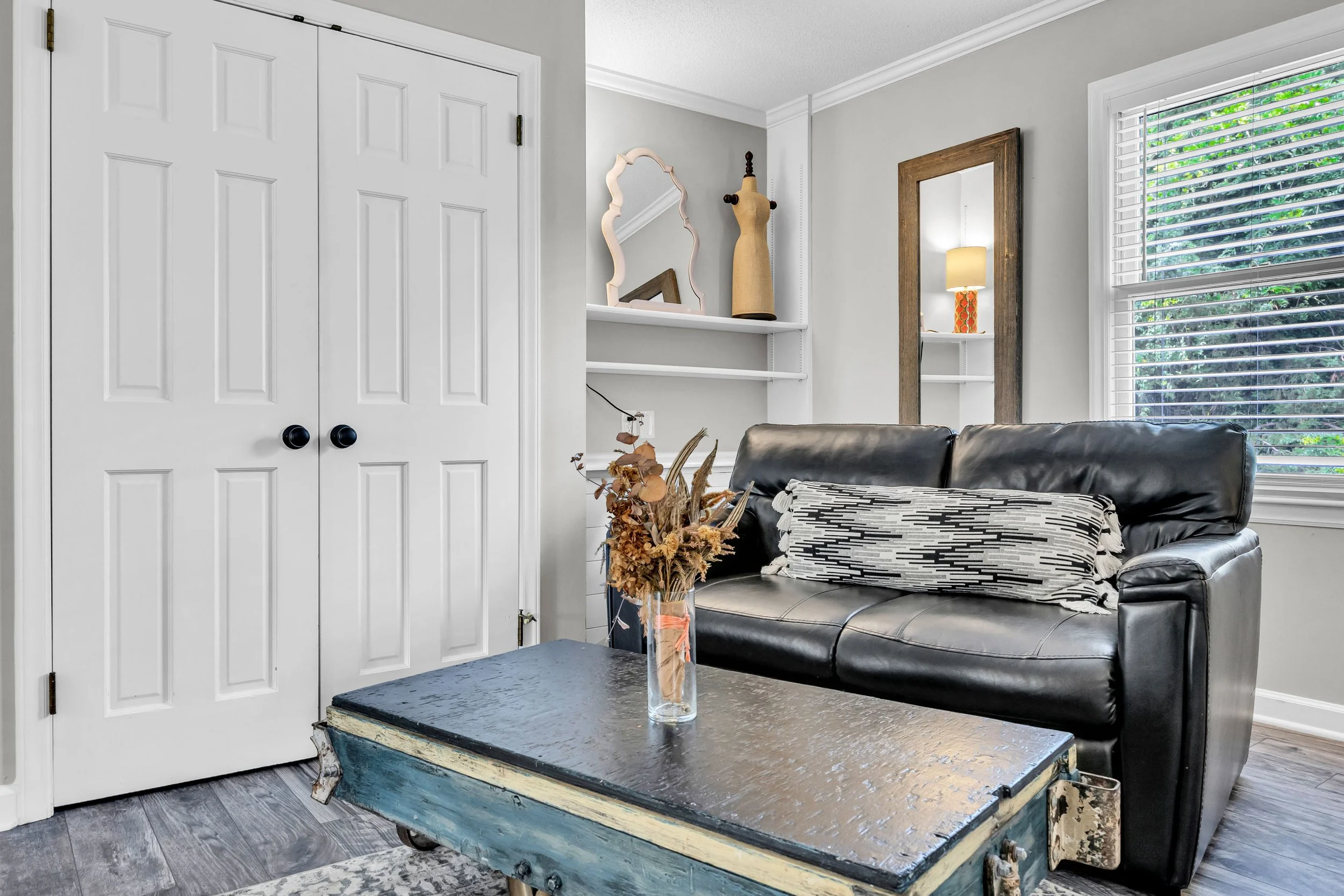 Living room with closet doors, a sofa with a patterned pillow, a rustic coffee table, a vase with dried flowers, and a shelf with decorative items and a mirror. Large window with blinds shows greenery outside.