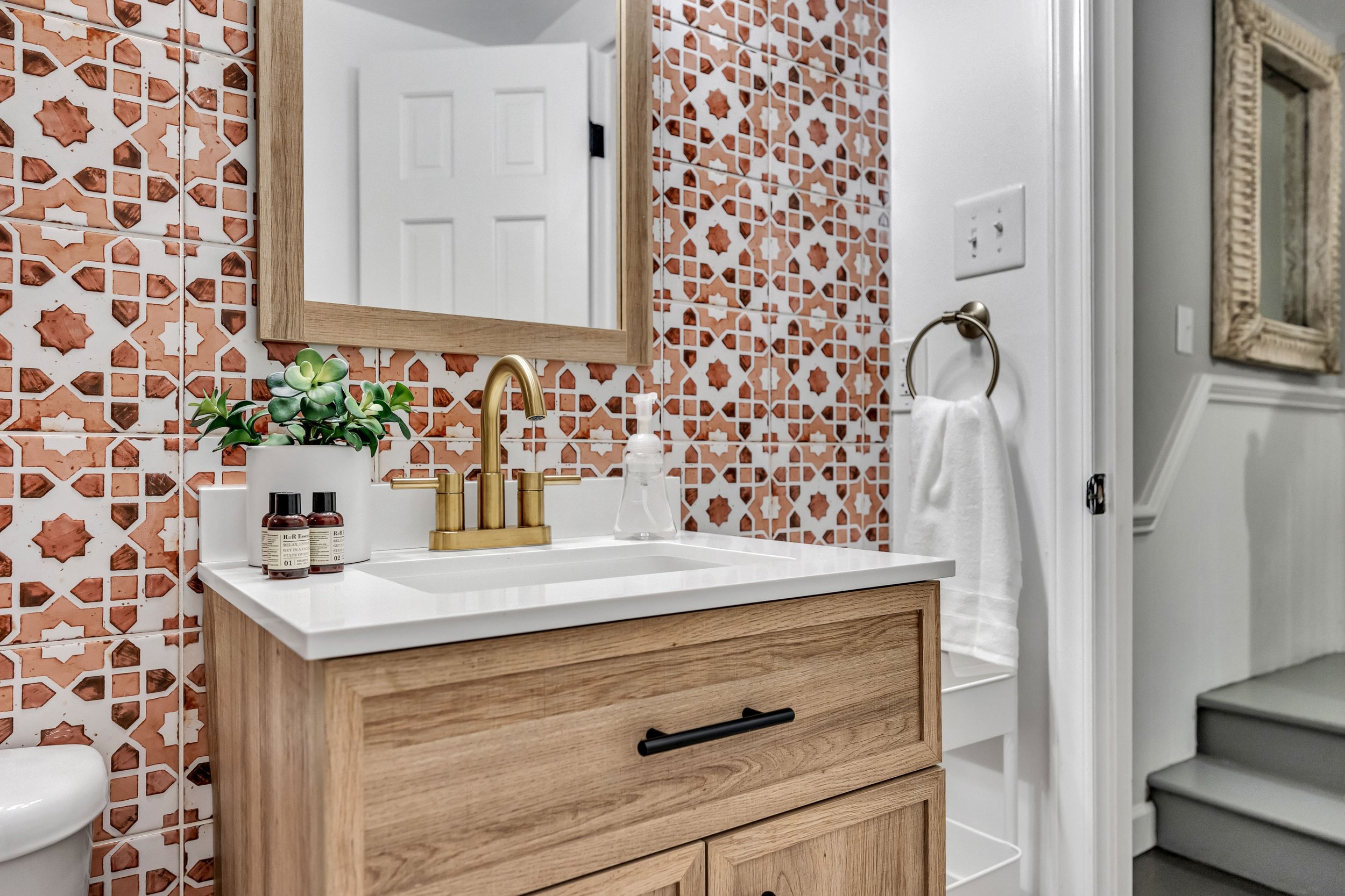 Bathroom with a wooden vanity, a mirror, a gold faucet, iodine bottles, a plant in a white pot, and decorative patterned tiles in shades of rust and white on the wall.