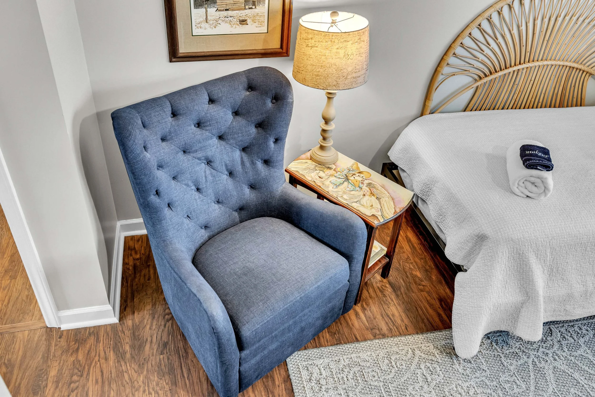 Interior of a bedroom featuring a blue upholstered armchair, a nightstand with a decorative butterfly design, a beige table lamp, a bed with a headboard made of rattan, and a rolled towel on the bed. A framed picture hangs on the white wall above the chair.