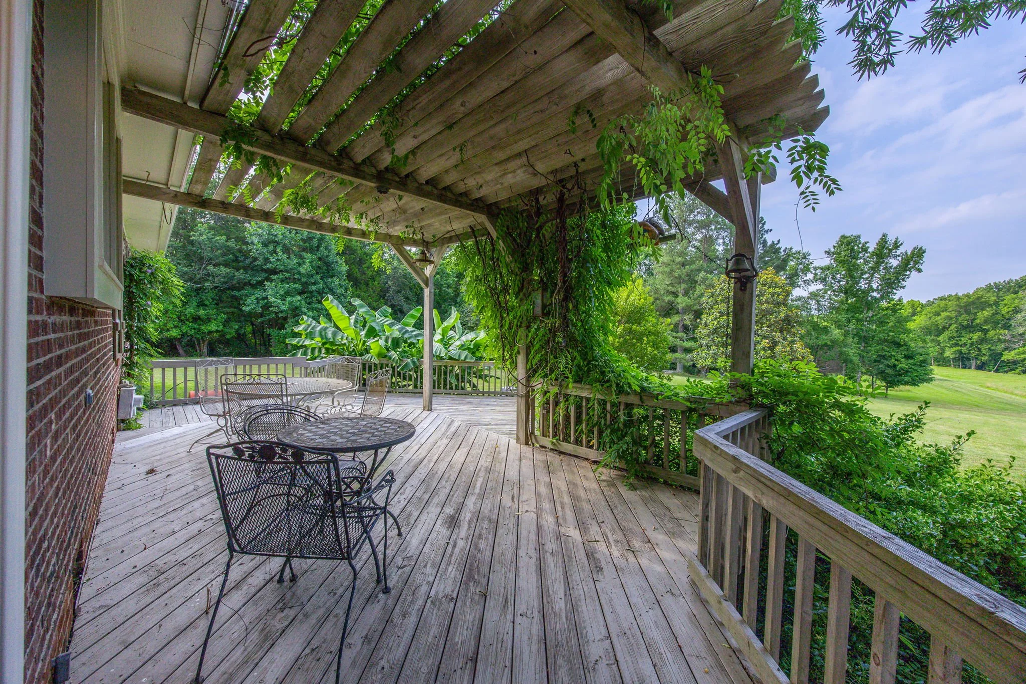 A wooden deck with metal outdoor furniture, surrounded by lush greenery and trees, with a sky overhead.