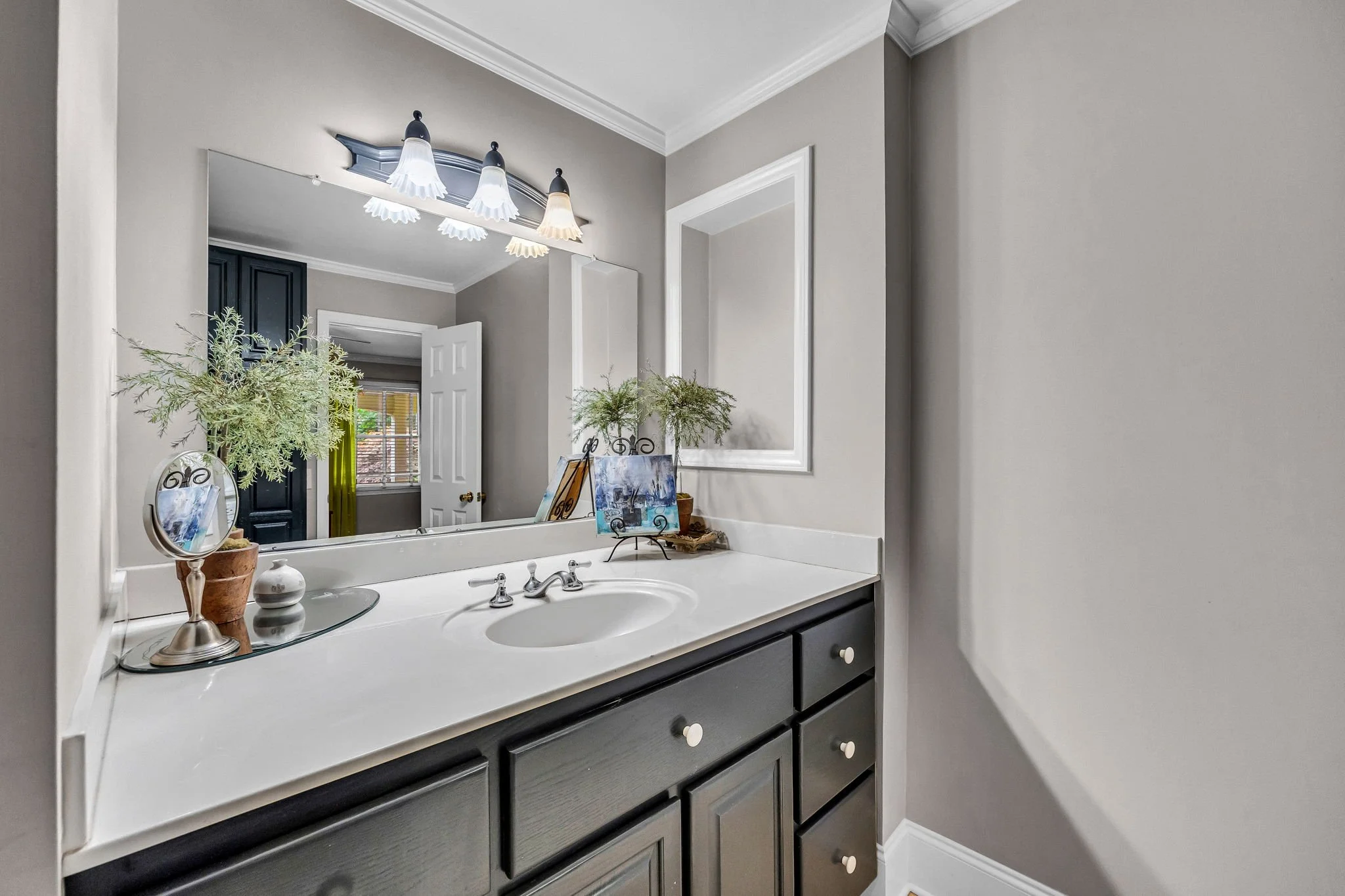 A bathroom vanity with a mirror, decorated with plants, a small clock, and a painting, with a three-light fixture above, reflecting a window with blinds and a door.