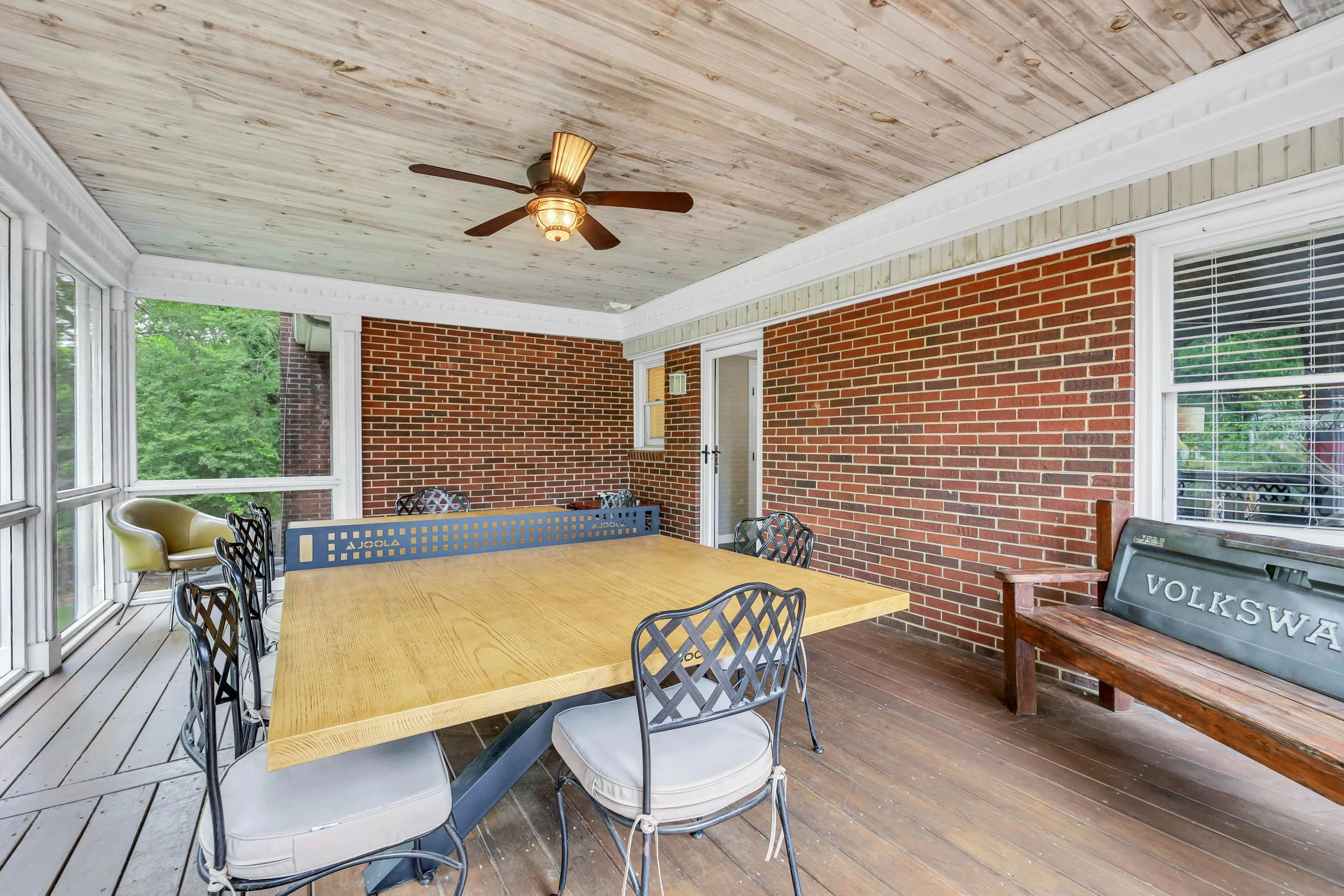 Enclosed porch with wooden ceiling, ceiling fan, brick walls, large windows, outdoor furniture, and a wooden bench with 'Volkswagen' painted on it.