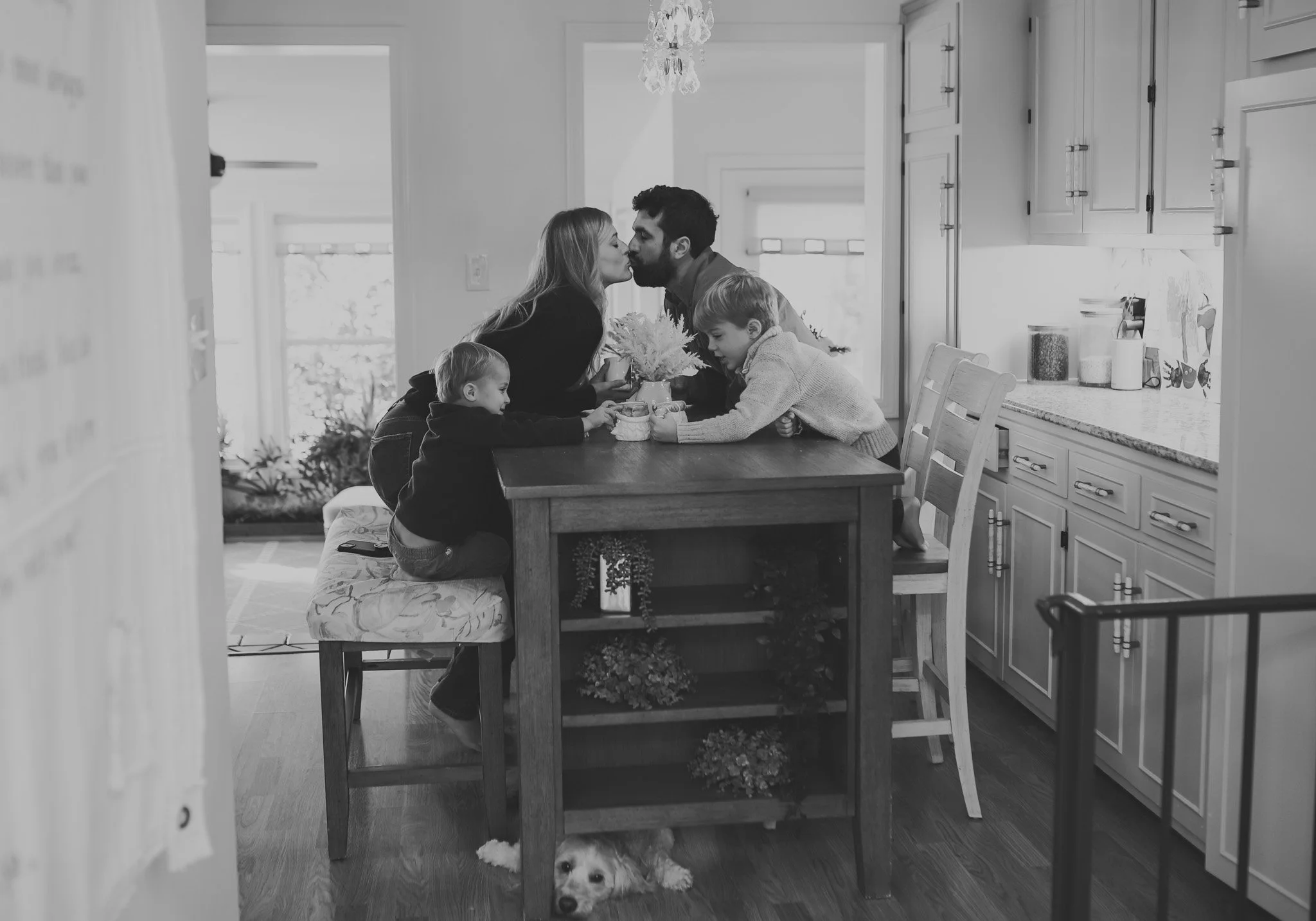 A black-and-white photo of a family in a kitchen. Two adults are leaning over a table, about to kiss. Two children are sitting at the table, one girl and one boy, with the girl holding a jar. There is a puppy lying on the floor under the table.