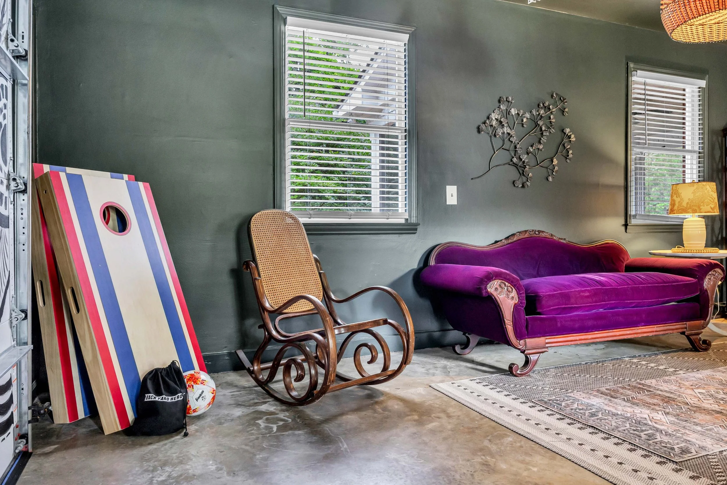 A garage with two windows, a purple velvet vintage couch, two wooden chairs, a metal wall art piece, a lamp, a striped cornhole board, a sports ball, a small bag, and a patterned rug on a concrete floor.