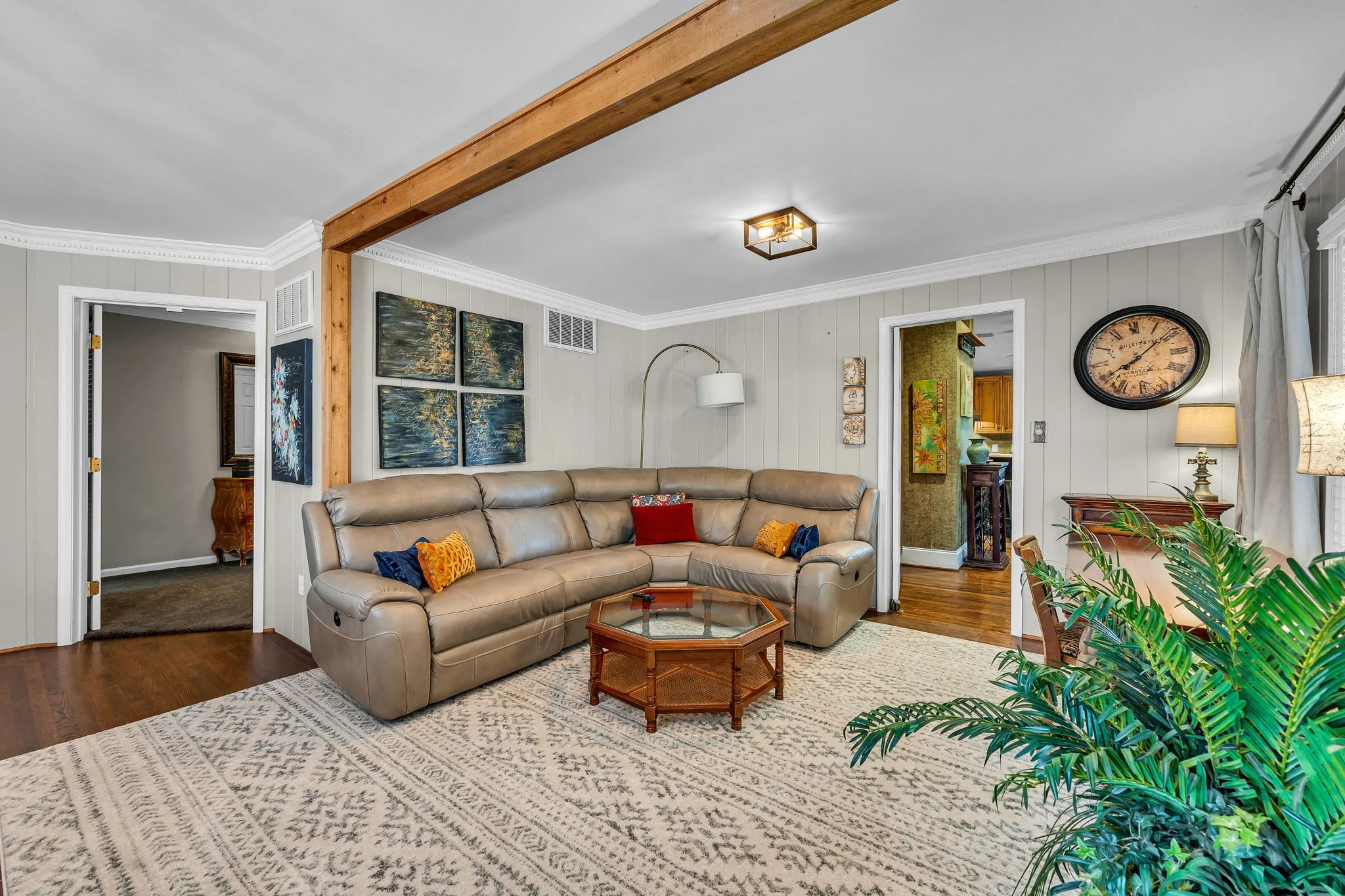 Living room with beige leather sofa, artwork on the walls, a wooden coffee table, and a patterned rug, with a wooden beam across the ceiling and a leafy plant in the corner.