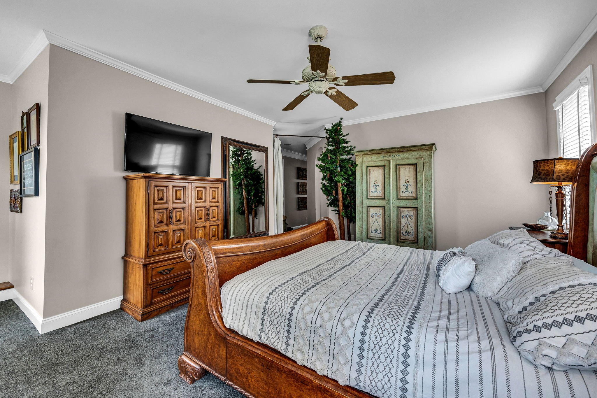 A bedroom with a wooden bed and striped comforter, a wooden dresser, a vintage green armoire, a wall-mounted TV, a mirror, a nightstand with a lamp, and a Christmas tree in the corner near a window with blinds.