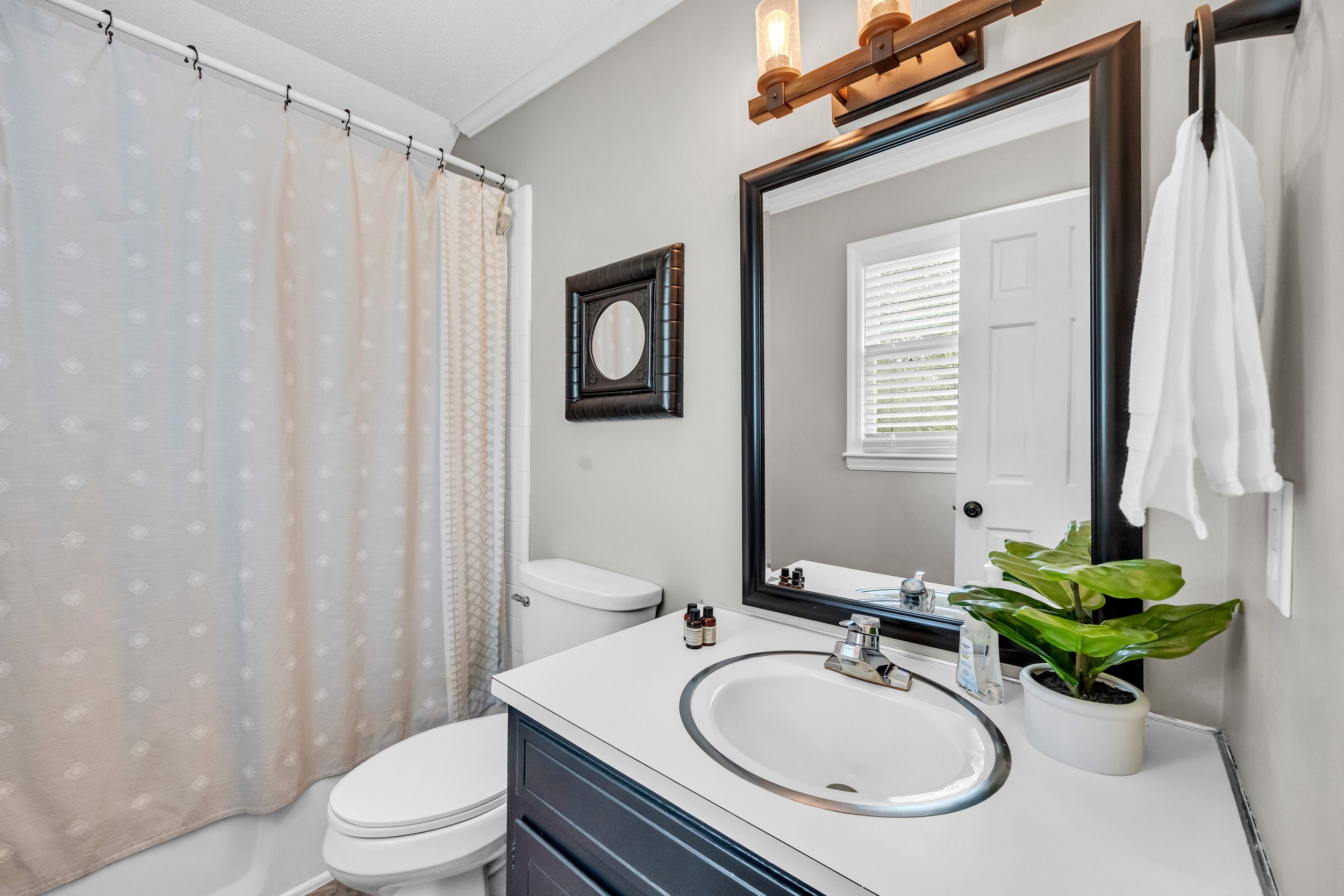 A bathroom with a white toilet, a white sink with a dark blue cabinet, a large mirror, a window with blinds, a potted plant on the sink, and bottles on the counter. A framed mirror is hung on the wall. A shower curtain with a subtle dotted pattern is on the left side.