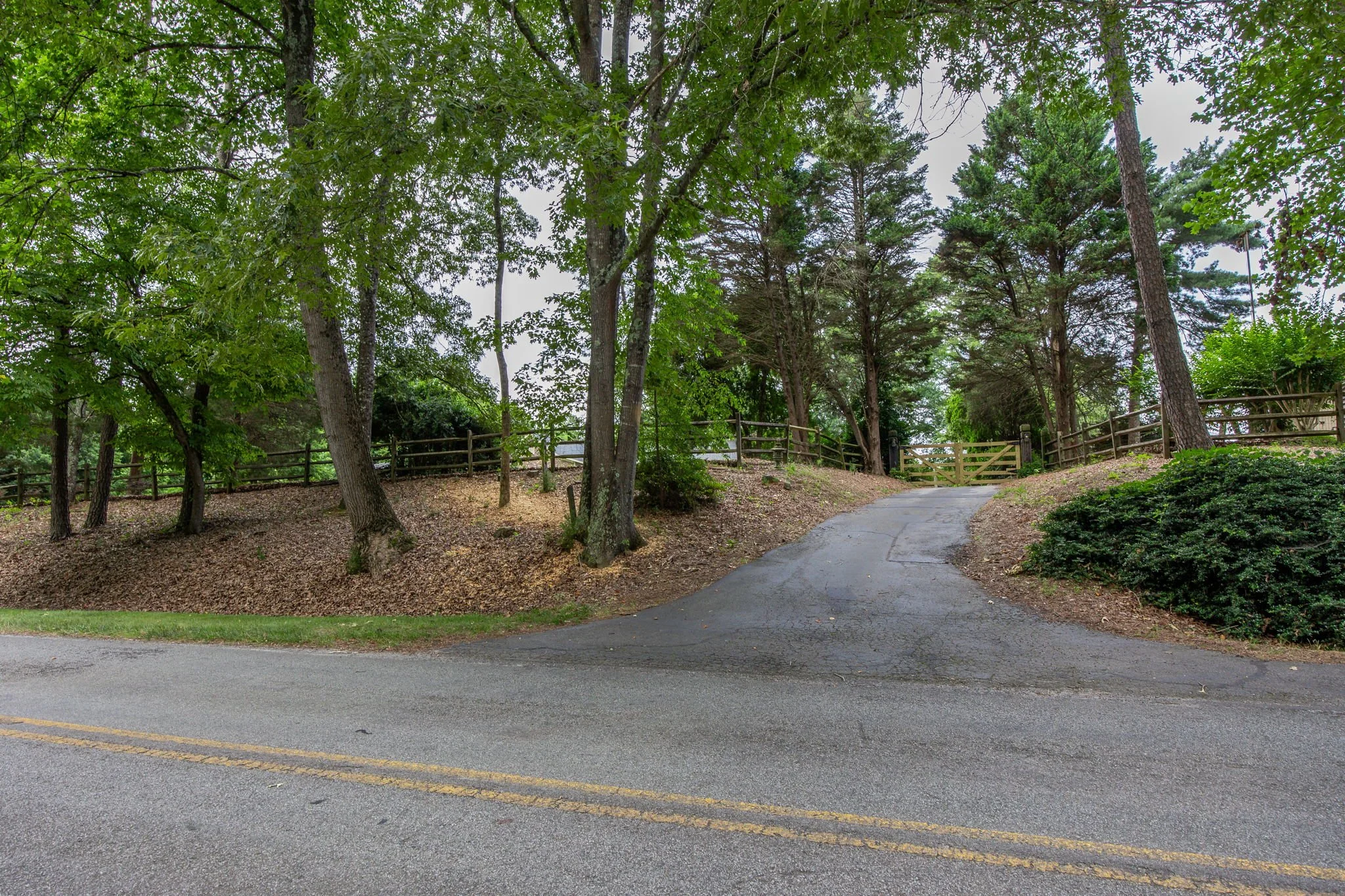 A paved road with a double yellow line, a sidewalk, and a wooded area with trees, a fence, and a gate.