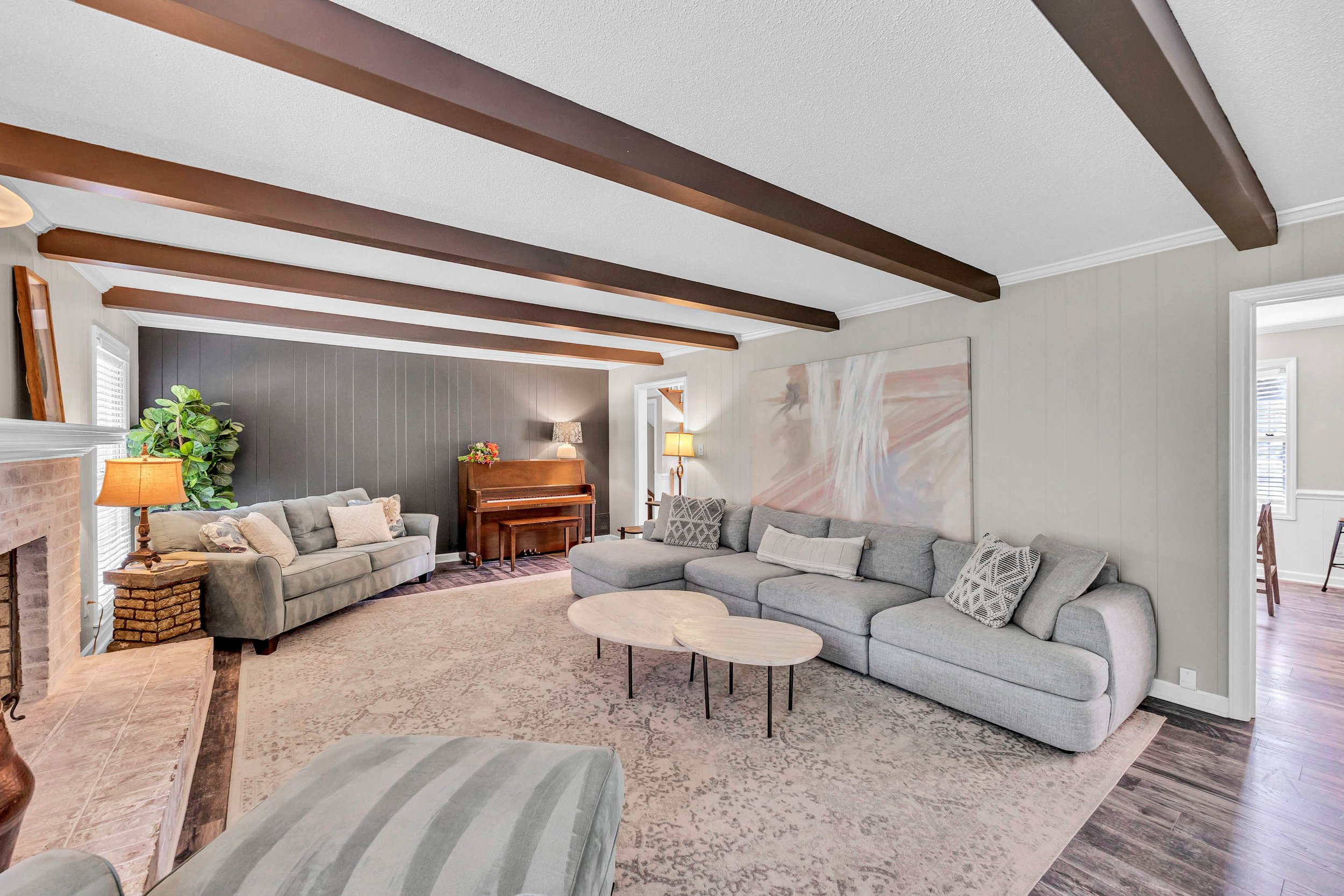 Living room with beige and gray couches, a wooden piano, and a large abstract painting on the wall. The ceiling has dark wooden beams, and there are two windows with white blinds, a side table with a lamp, and decorative pillows on the couches.