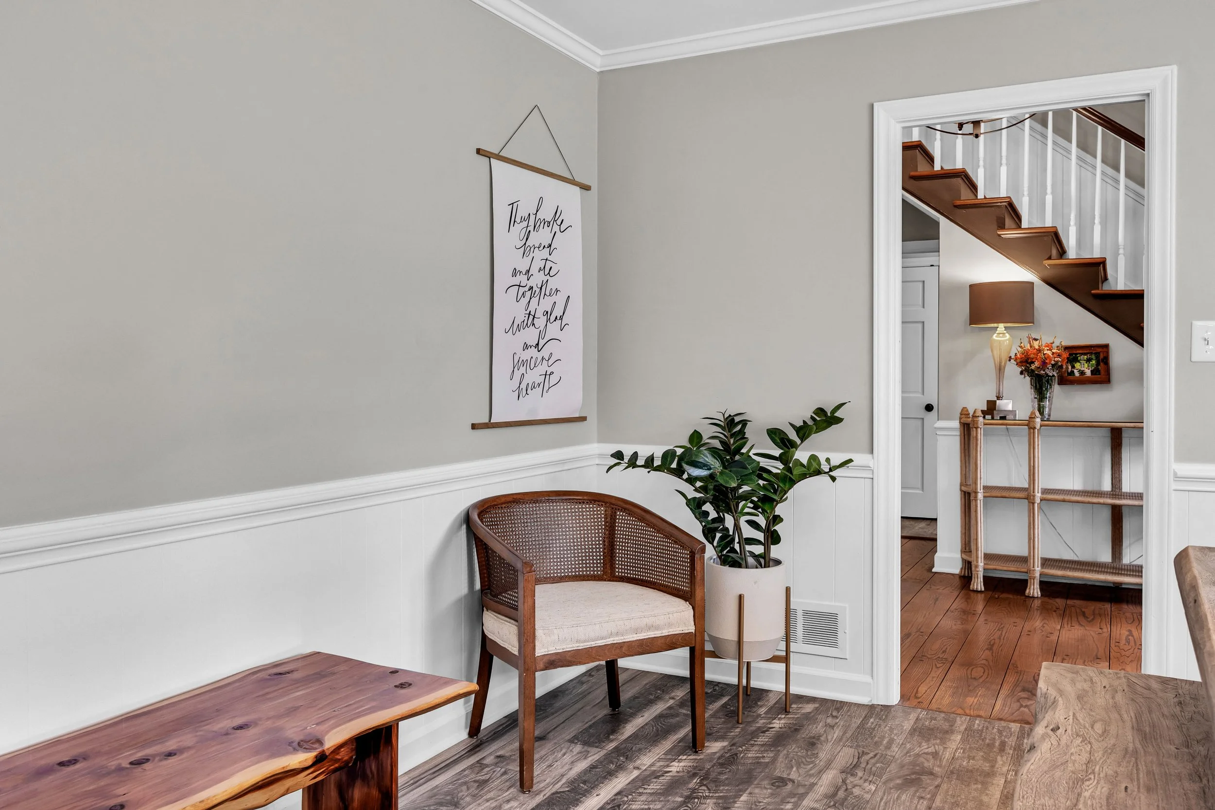 Interior of a home with beige walls, a white wall molding, and a staircase with wooden steps and white spindles. There is a wall banner with cursive text, a wooden chair with a cushion, a green potted plant, a table, a floor vent, and decorative items including a lamp with a brown shade and a vase of flowers.