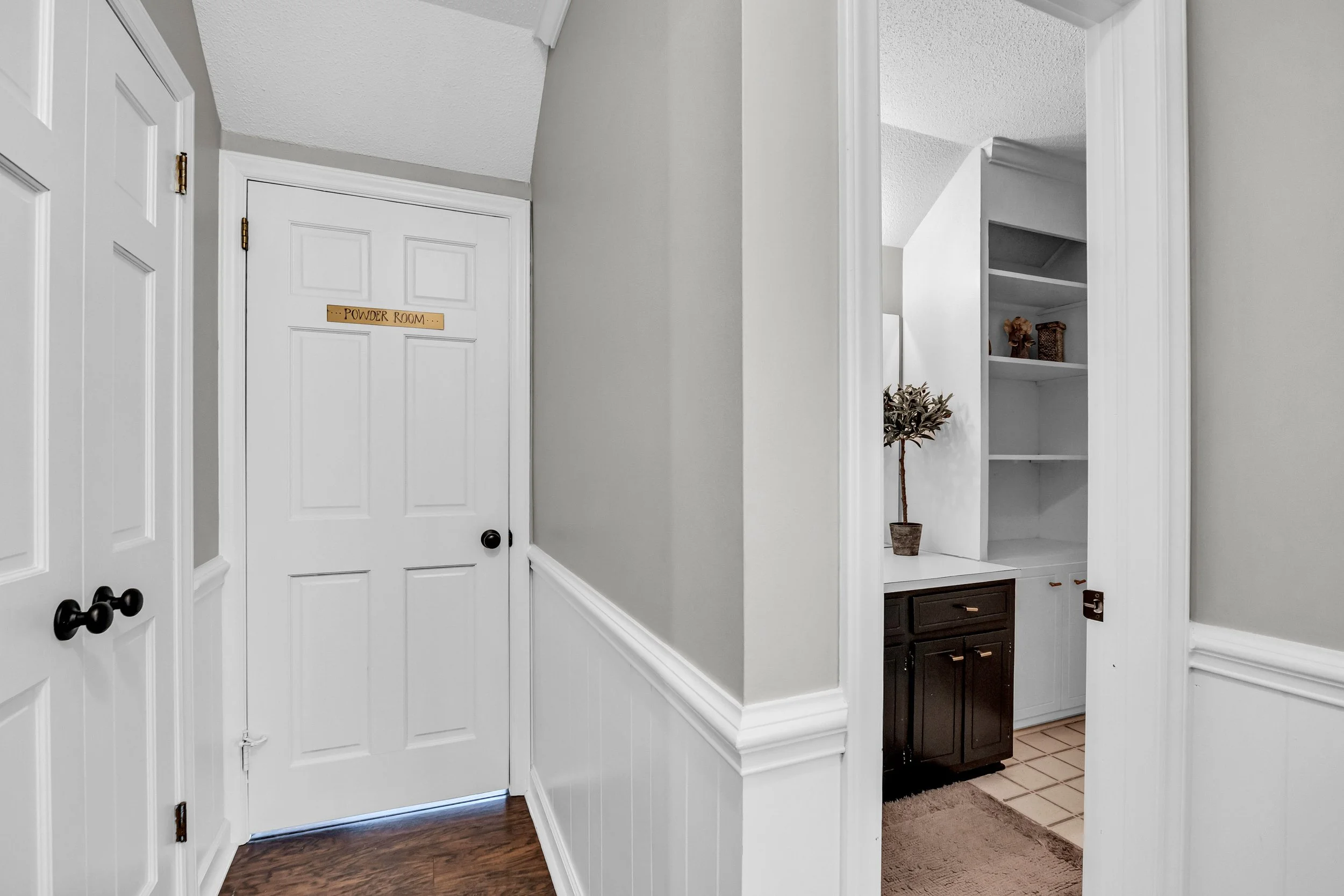 Interior view of a house, showing a hallway with white walls and doors, and an opening leading to a stairway and a small bathroom or laundry area with dark cabinets and a potted plant.