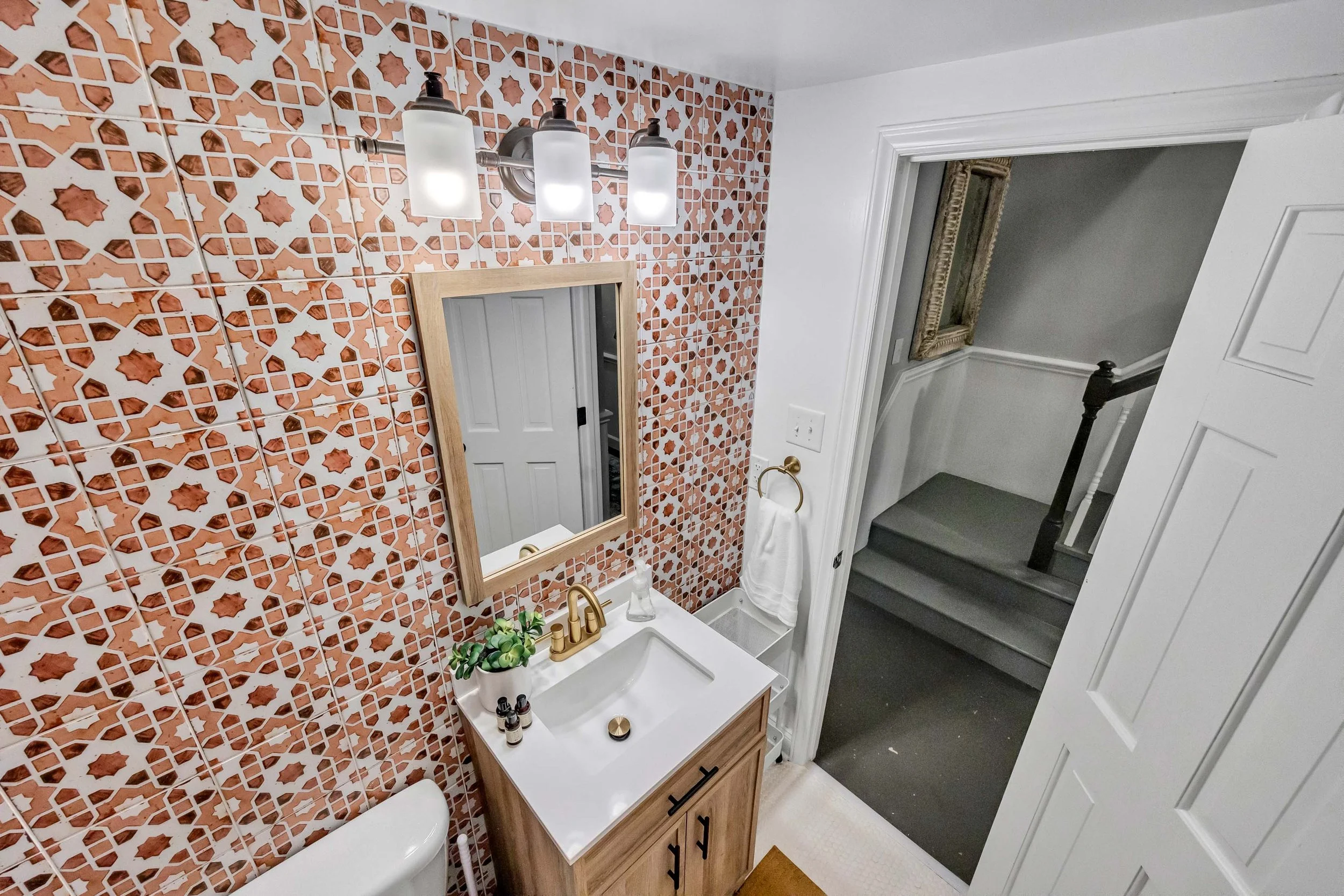 A bathroom with a vanity, mirror, and light fixture. The wall has decorative tiles with a geometric pattern in reddish-brown and white. A small potted plant and toiletries are on the vanity. There are stairs and a doorway in the background.