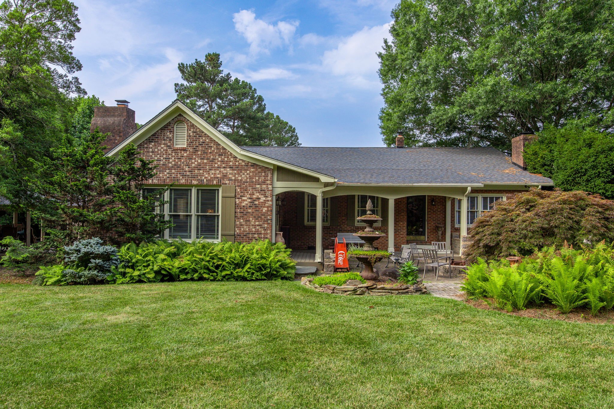 Red brick house with a covered porch, surrounded by lush green trees and shrubs, with a well-maintained lawn and outdoor seating area, under a partly cloudy sky.