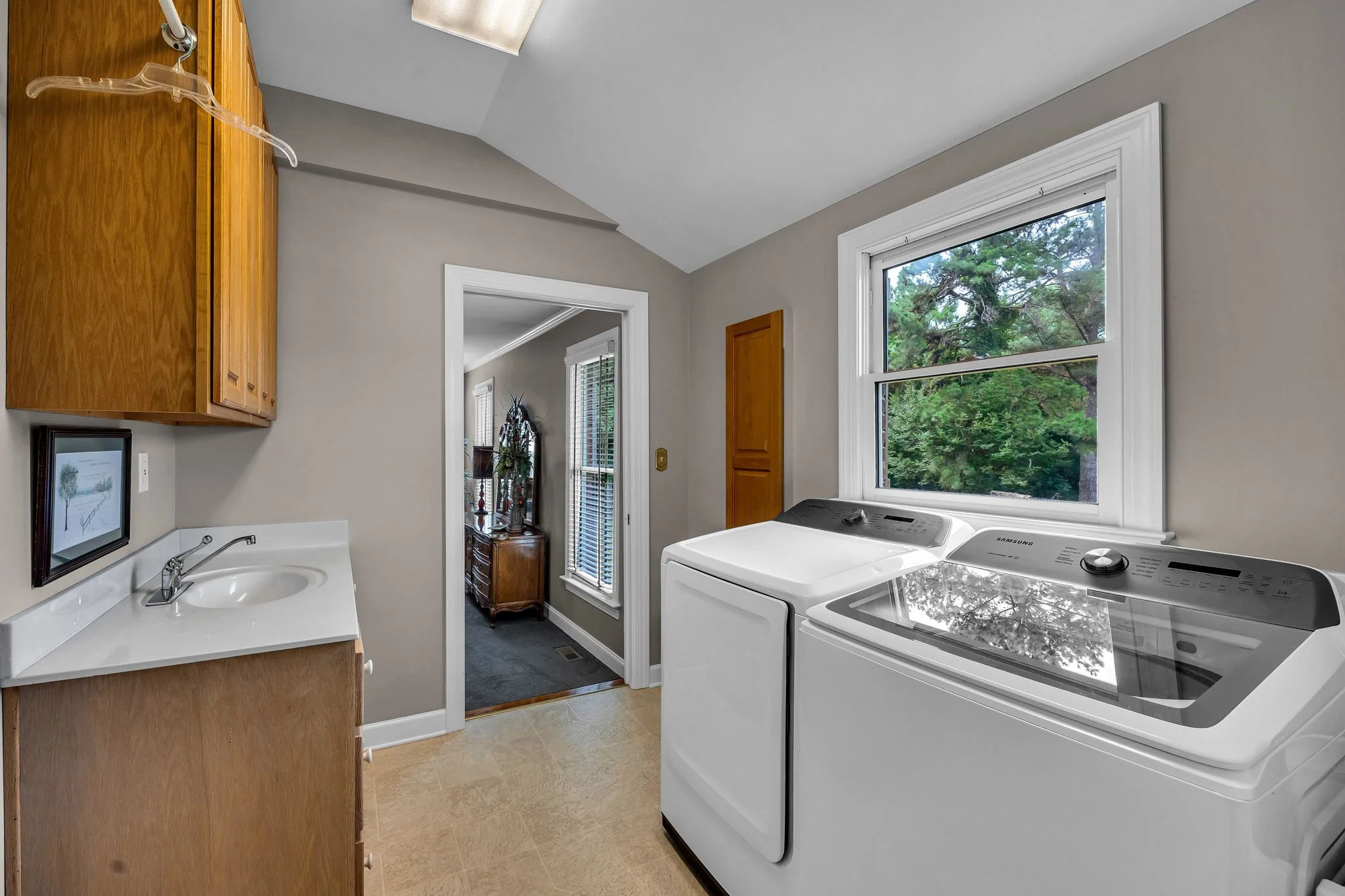Laundry room with white washing machine and dryer, a window showing green trees outside, wooden cabinets, a small sink, and a doorway leading to another room.