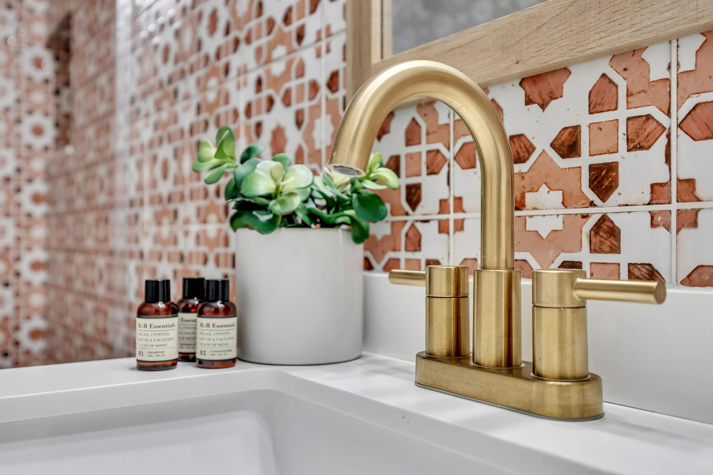 A bathroom sink with a gold faucet, a small potted succulent, and three small bottles of skincare products on a white countertop. The background features a decorative red and white patterned tile wall and a wooden shelf.